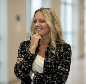 Alex Colley, a smiling woman with long blonde hair in a black and white patterned blazer, standing in an indoor setting.