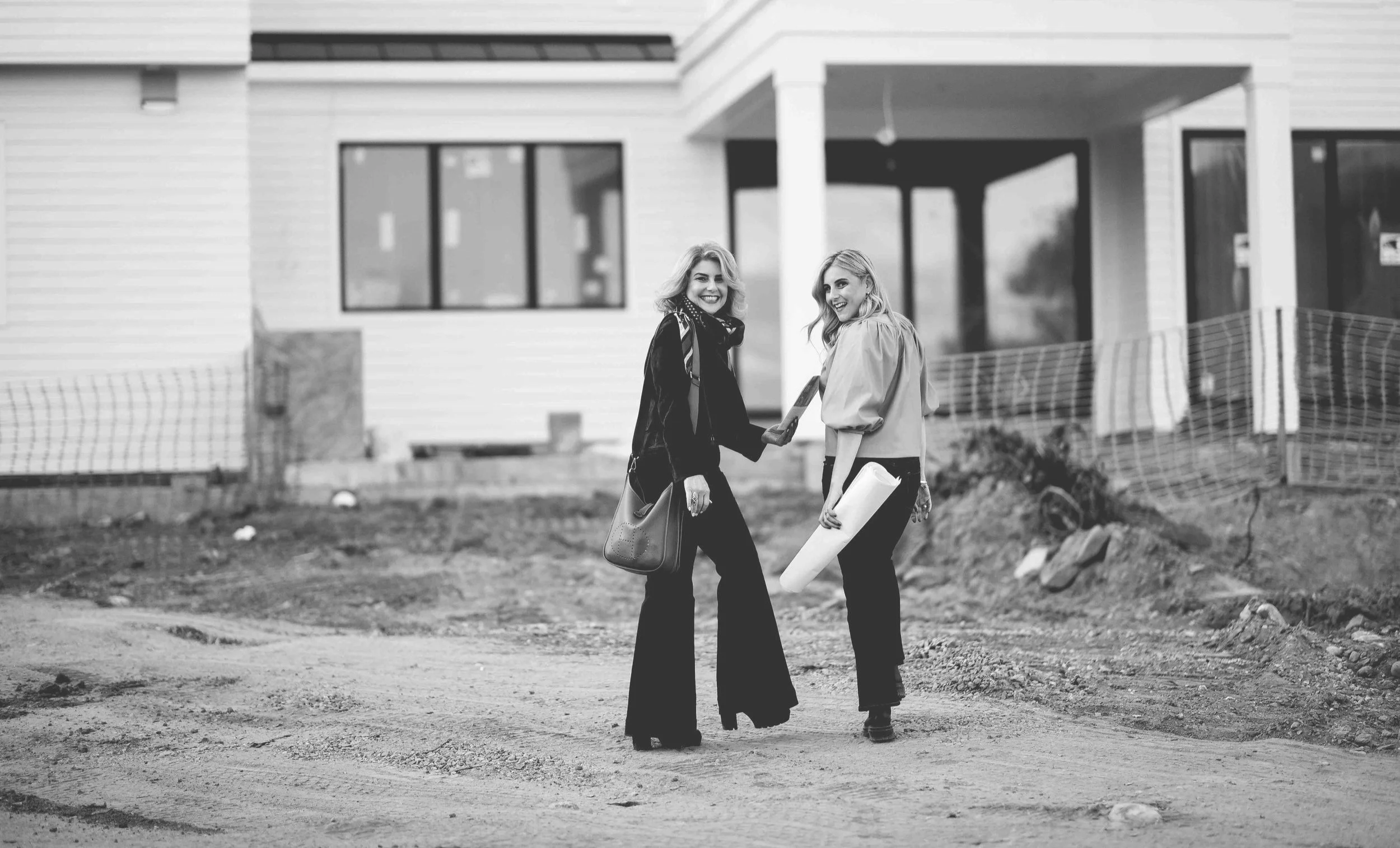 Two women smiling, holding hands in front of a modern white house under construction, one with a blueprint.