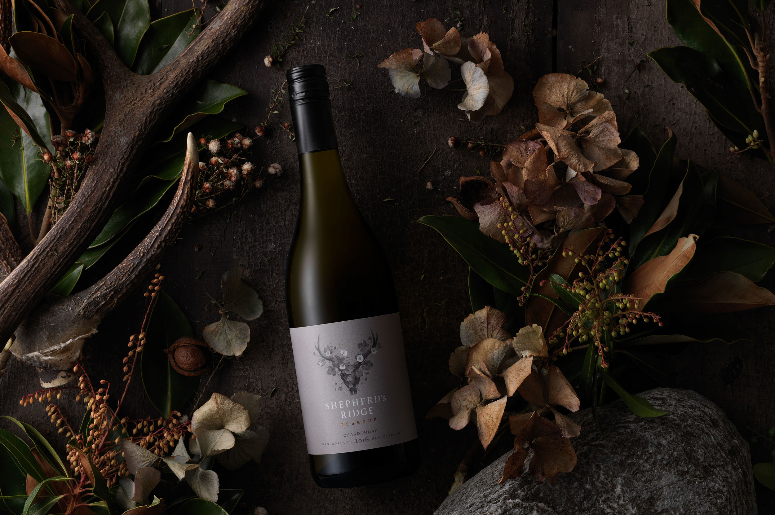 However rather than featuring the traditional geographical features of the Marlborough region or a cliched reference to a shepherd, we decided to create a much more abstract interpretation that evoked the origins and culture of the wine. Animal skulls can be found adorning many South Island houses and cribs, but they are more than ornaments-they are symbols of respect for the creatures that have often sustained life and worked in harmony with the land. To Curious, this was a perfect analogy for the Shepherd’s Ridge vineyards. A sense of regeneration was then also introduced to build in an extra layer of story telling. This was achieved by elegantly interlacing natural vegetation around the iconic skull shape. Anton Petrov from Watermark Creative captured our vision perfectly with a beautifully understated illustration. It’s simple black and white colour palette and strong sense of structure creates a very powerful image base-but this was then delicately balanced with the flowing cursive lines of vines, leaves and flowers. To complement this iconography, we chose to incorporate subtle warm grey hues and copper foil to add premium cues and a discreet pop of colour. The typography has also been sensitively handled to enhance the sophistication of the label. Once the primary packaging had been established, we then moved on to creating a suite of imagery that could be used for all forms of media. Given that Shepherd’s Ridge Reserve is a totally new brand, it was important to feature the bottle and label-as this would allow the consumer to create a strong visual connection at point of purchase. However we also wanted to ensure that there was a symbiotic relationship between the packaging and its origins, so each varietal was surrounded by colour co-ordinated flora and natural stone. The subtle lighting and earthy tones then completed the narrative.