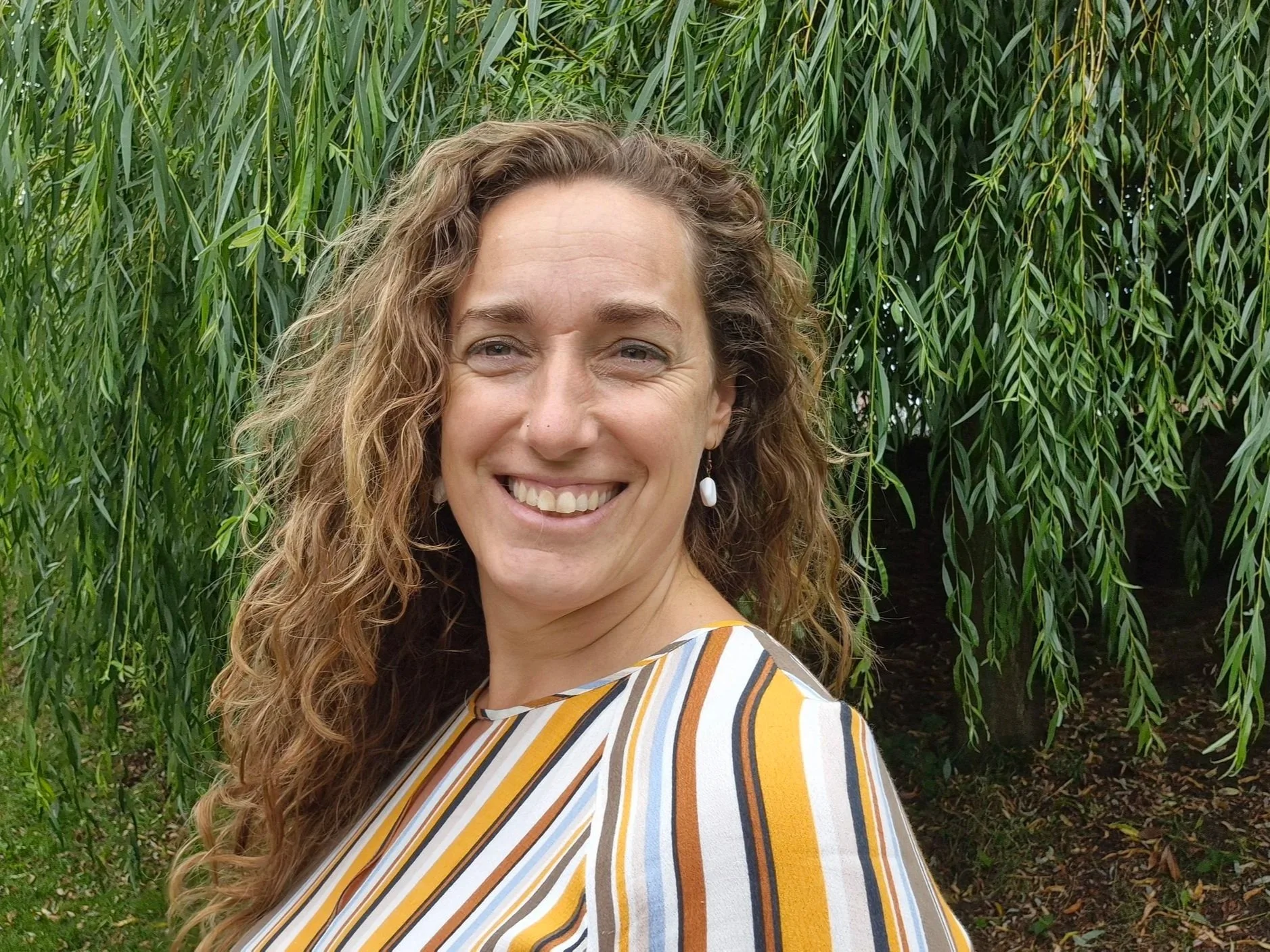 Vrouw met golvend bruin haar lachend in de buitenlucht, achtergrond met groene bomen en planten.