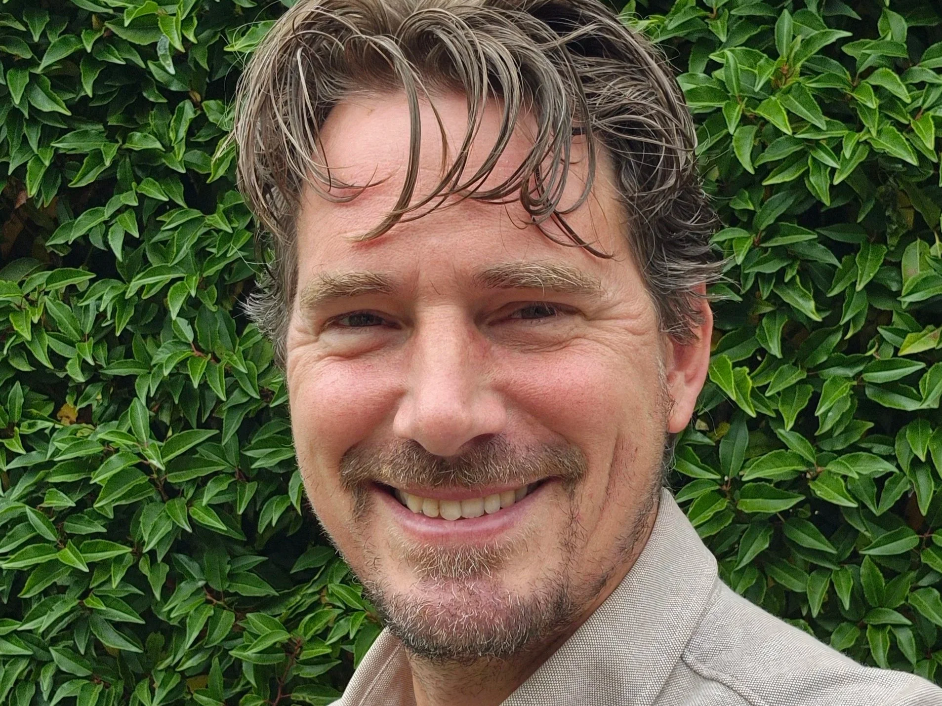 Mann met glimlach, kort grijs haar en een baard, lachend voor een groene struik.