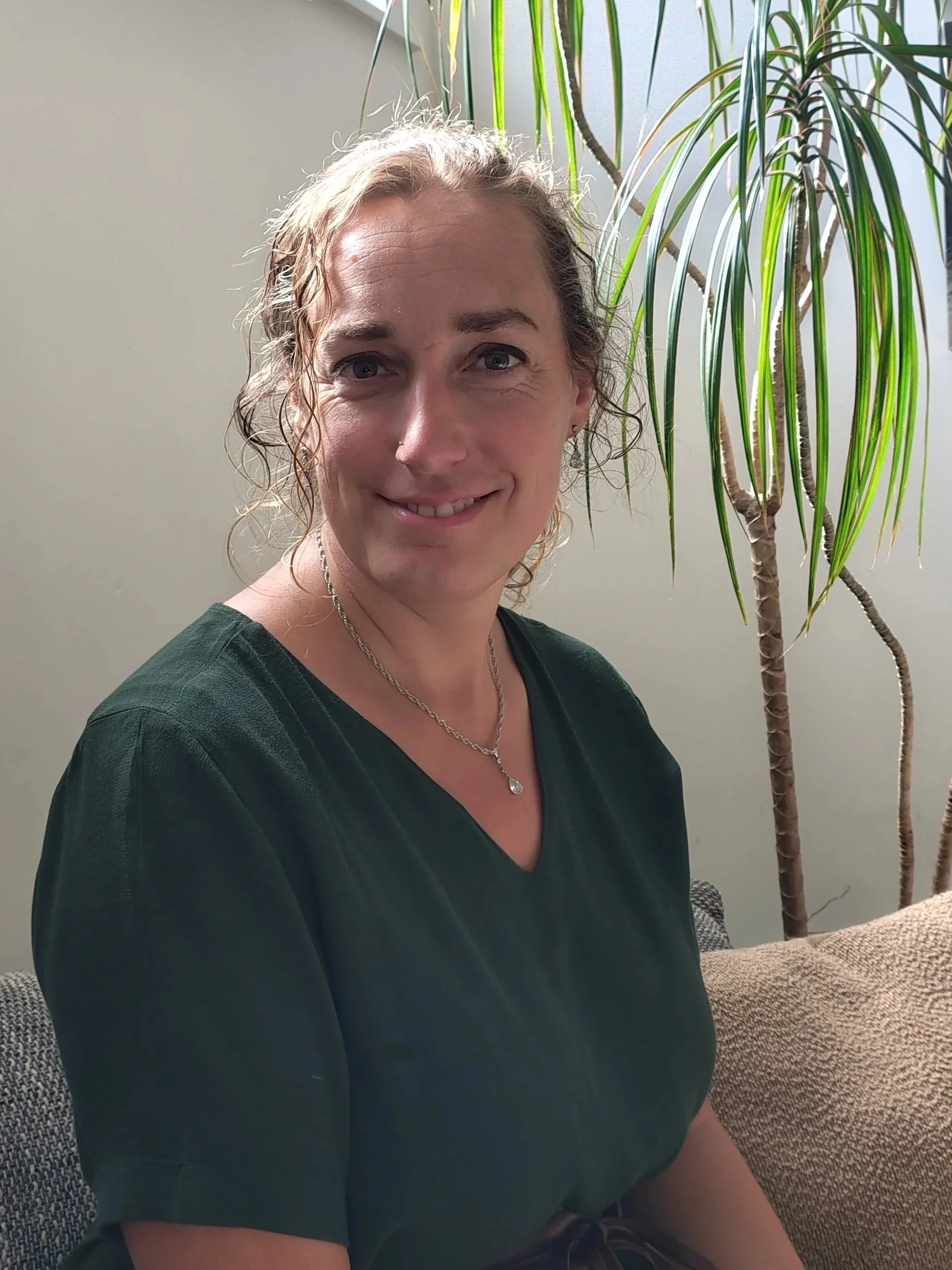 Portret van een glimlachende vrouw met blond, gekruld haar, zittend op een bank naast een grote plant, indoors.