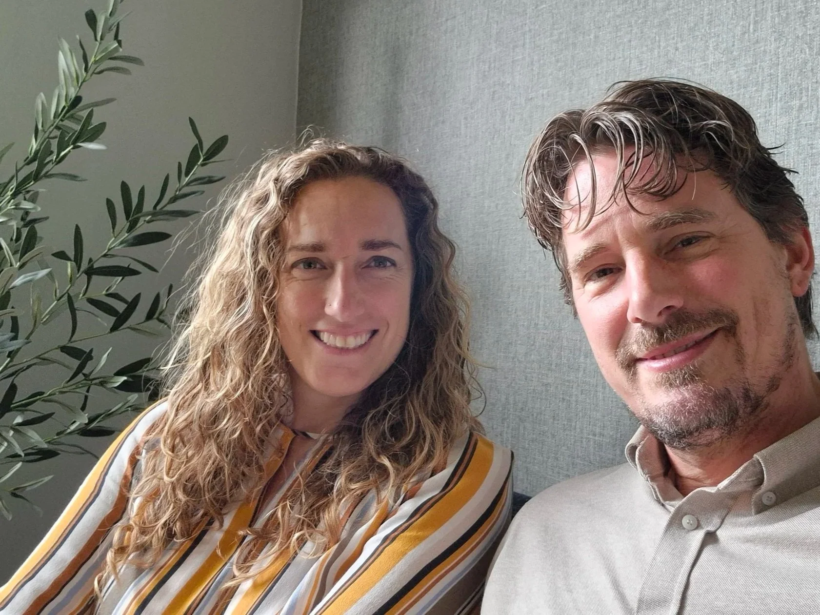 Een man en vrouw poseren voor een selfie, lachend, met een groen plant en een blauwe muur op de achtergrond.