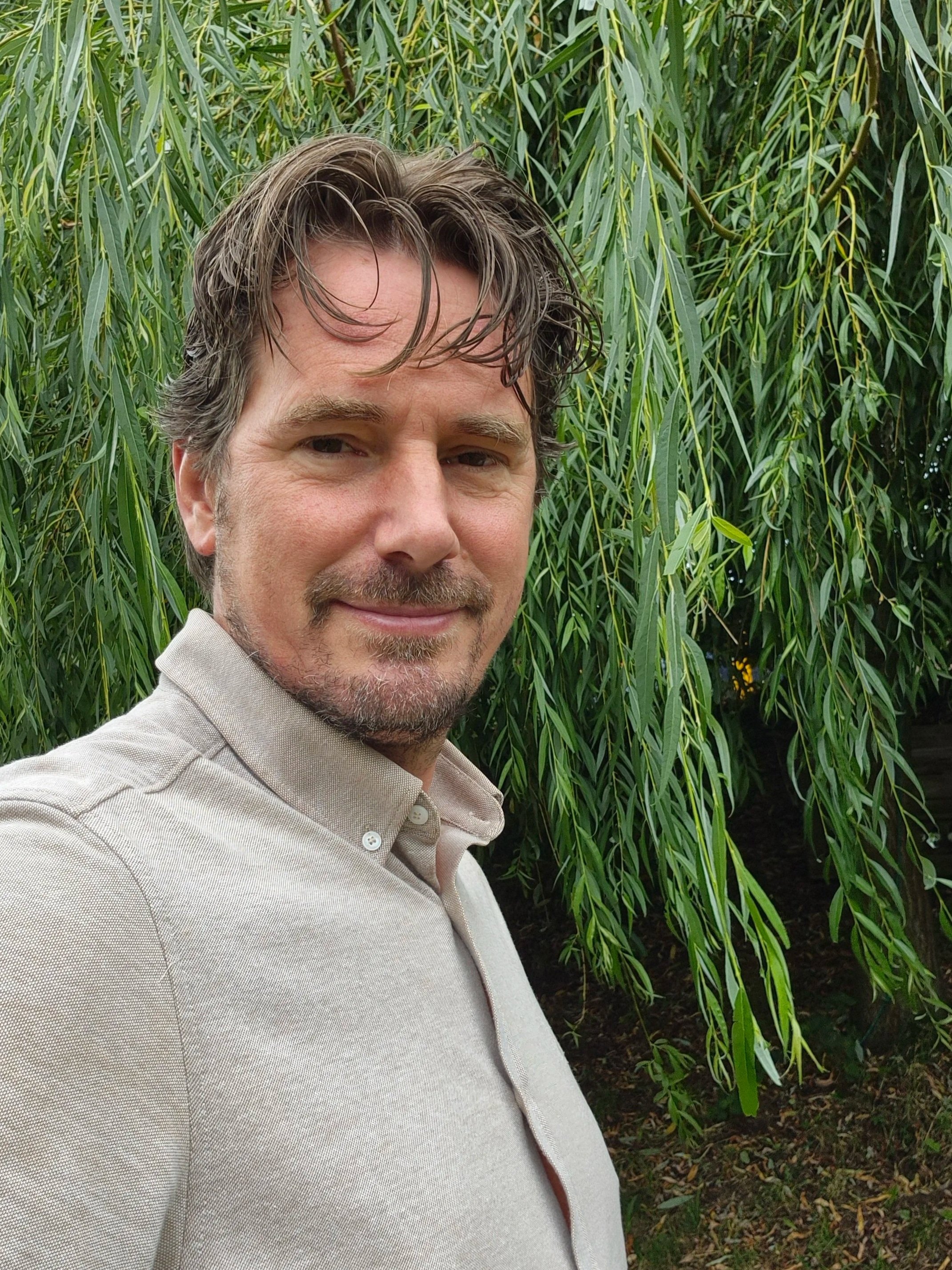 Een man met donkerbruin haar en een lichte huid die in een tuin staat, achter een grote boom met groene bladeren.