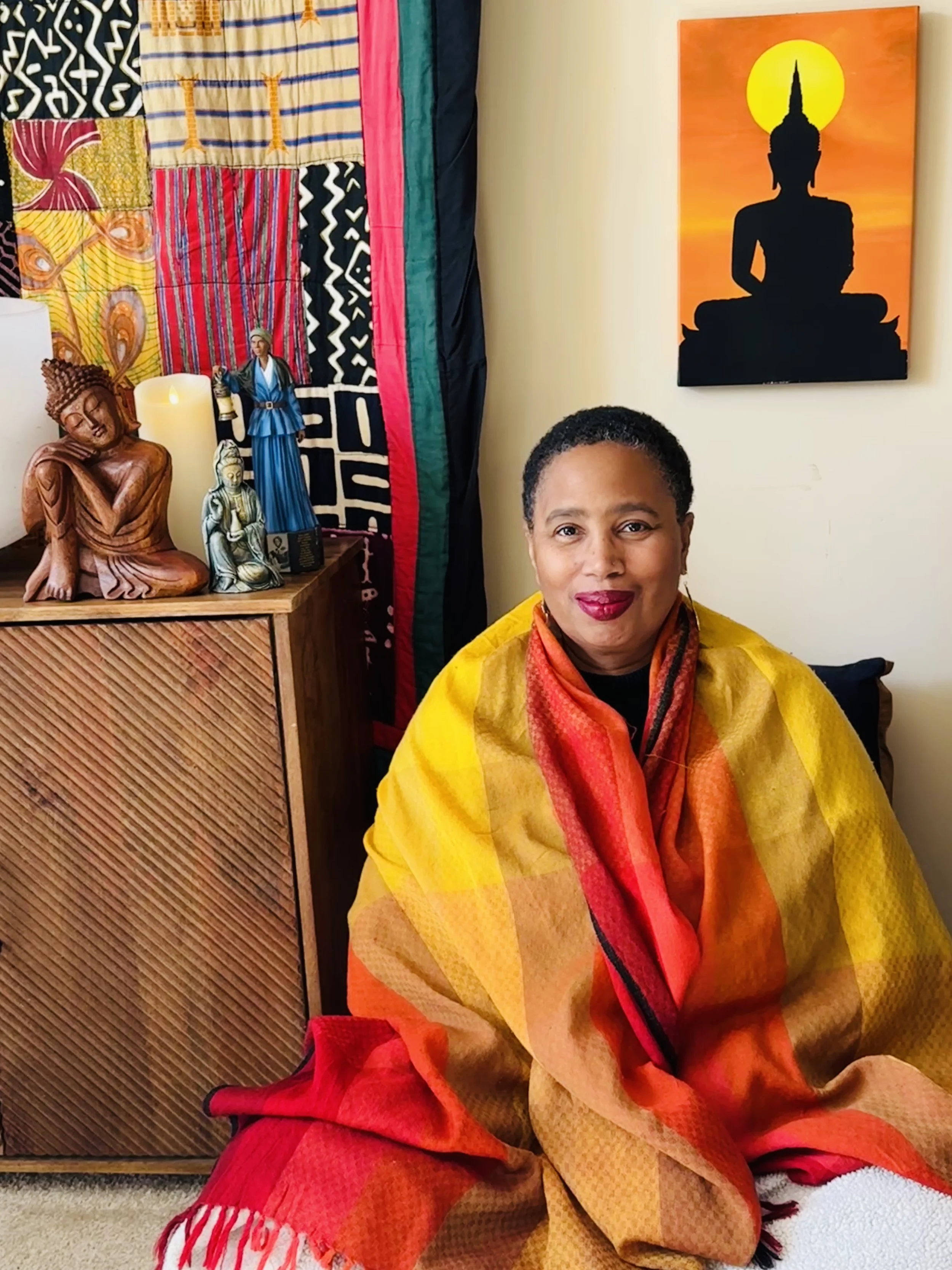 Aishah Shahidah Simmons seated on meditation cushions, wrapped in a yellow-orange shawl, with a silhouetted Buddha image on the wall behind her against an orange background with a sun or moon.