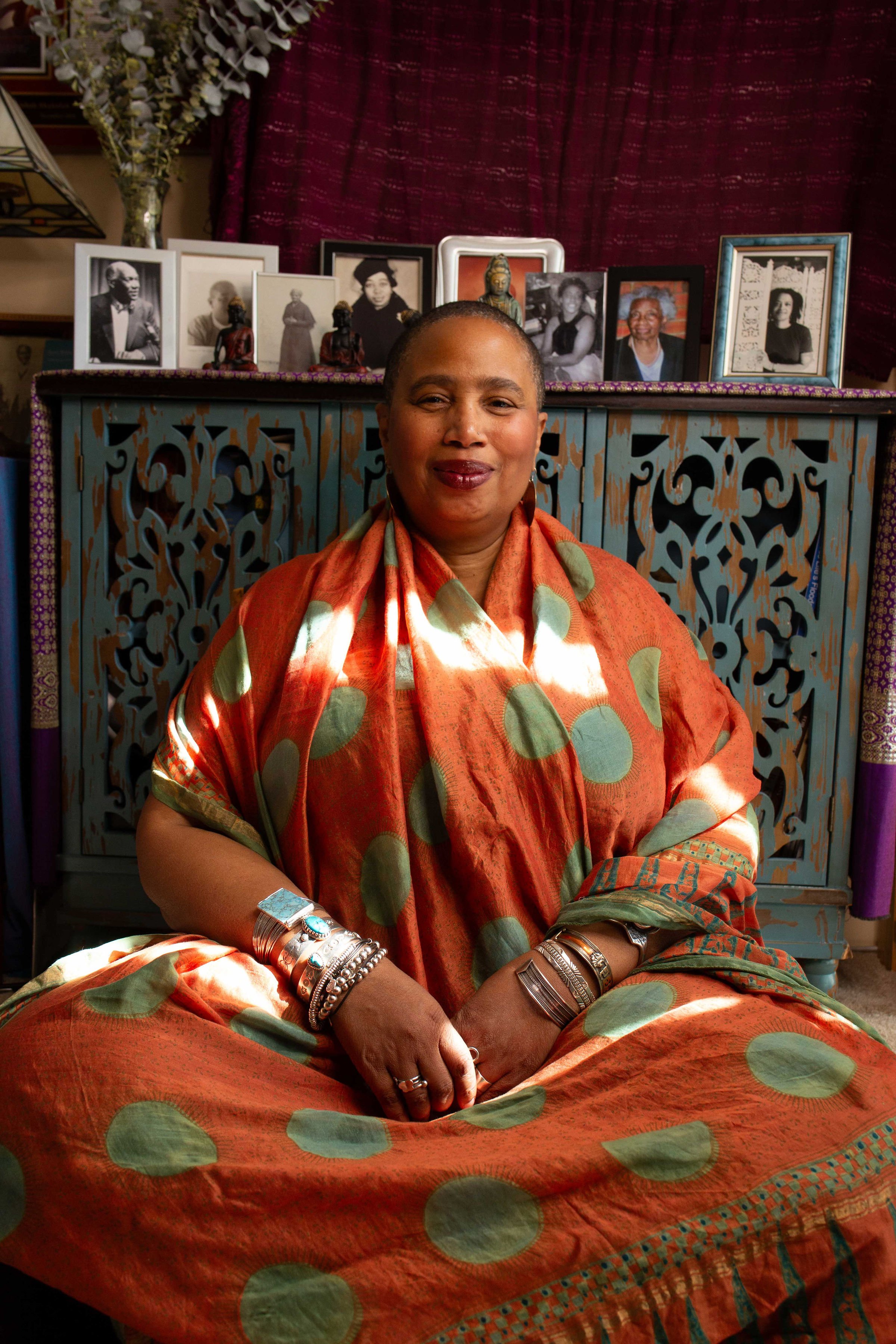 Aishah Shahidah Simmons seated before her ancestral altar, smiling gently, hands folded. Photo by Studio TSHAY