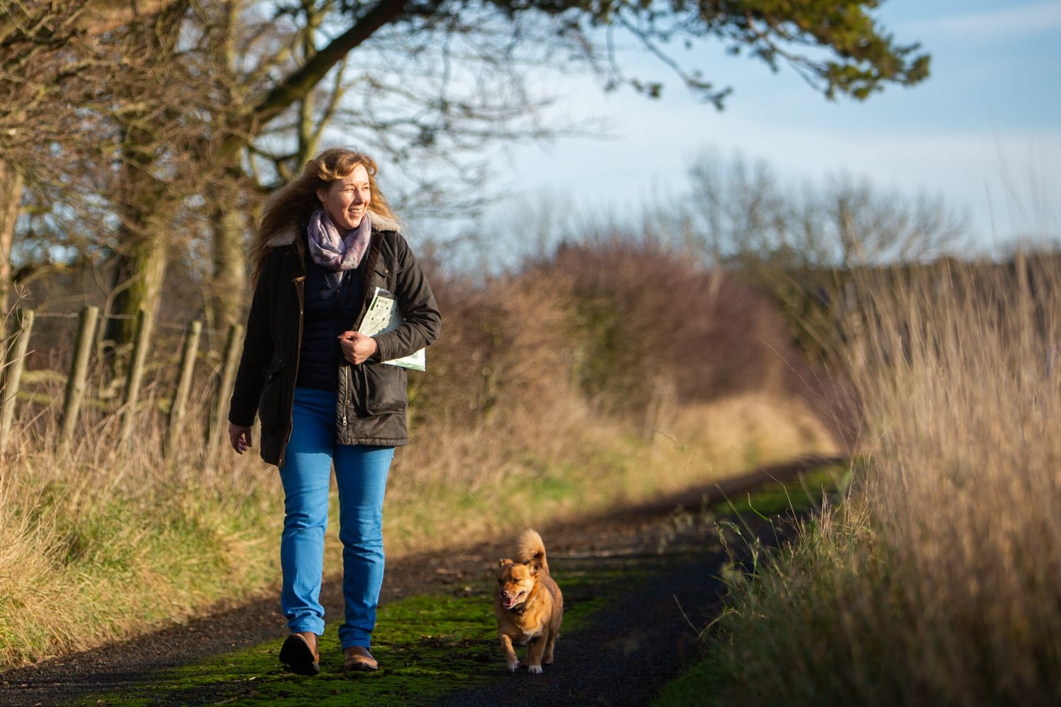 Anne Gray, a woman with big blonde hair, walking down a country path with a small dog business photographer northumberland