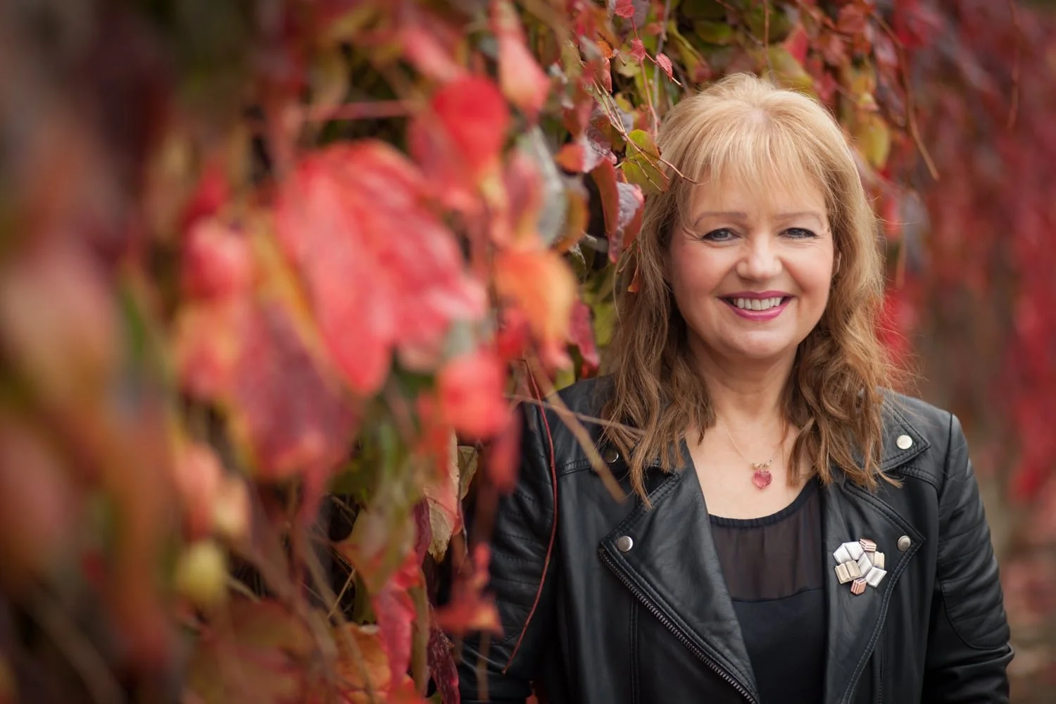 Woman in a black leather jacket smiling while leaning against a wall adorned with red and orange leaves in a range of colour by sarah jamieson headshot photographer northumberland