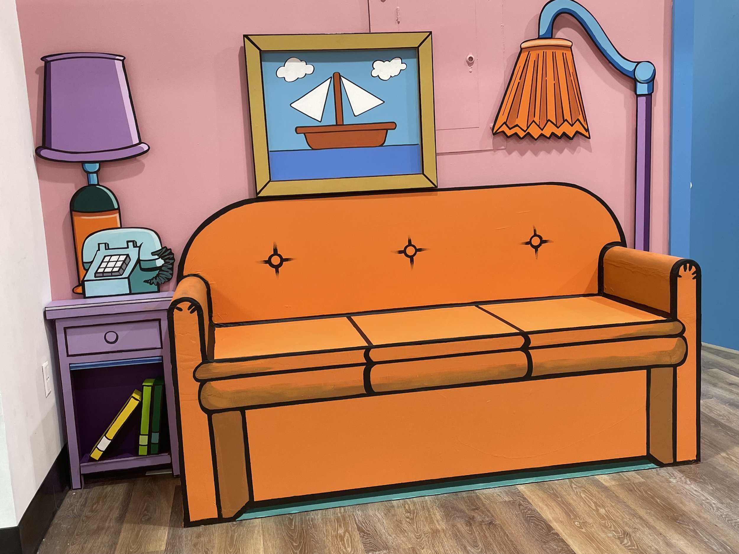 Colorful corner with a cartoon-style orange sofa, purple and orange lamps, a purple side table with a vintage rotary phone and books, and a framed picture of a sailboat on the wall.