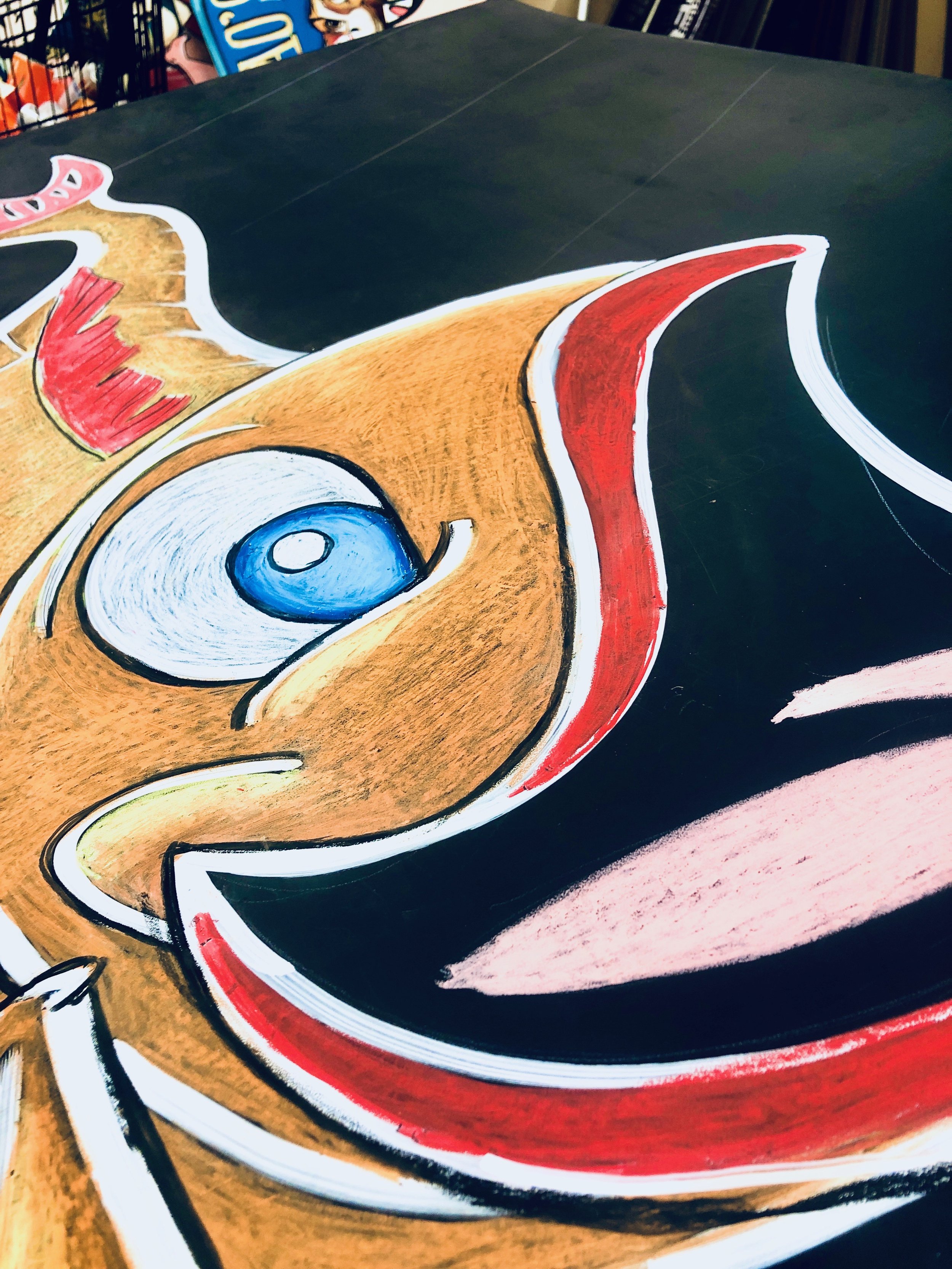 Close-up of a large, colorful chalk drawing on a black surface featuring a stylized cartoon koi fish with big blue eyes, a pink tongue, and red and white accents.