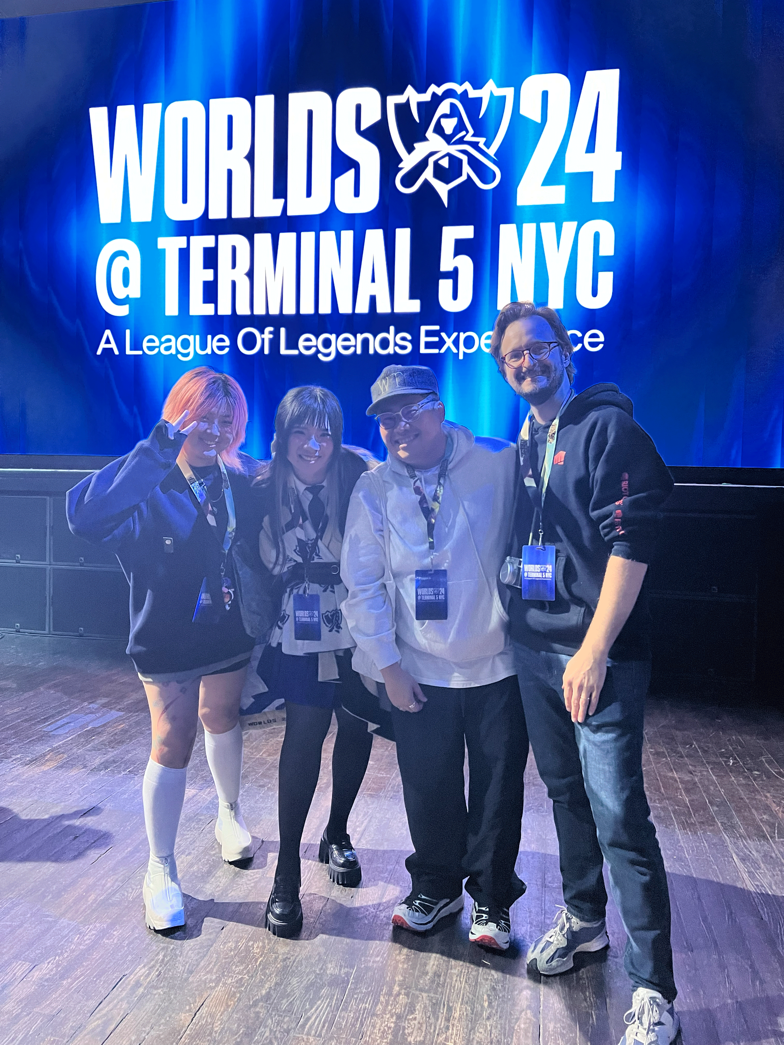 Four people posing together at a League of Legends esports event, with a large blue sign in the background reading "WORLDS 24 @ TERMINAL 5 NYC" and a League of Legends logo. The group includes two women and two men, all smiling and dressed casually, wearing event lanyards.