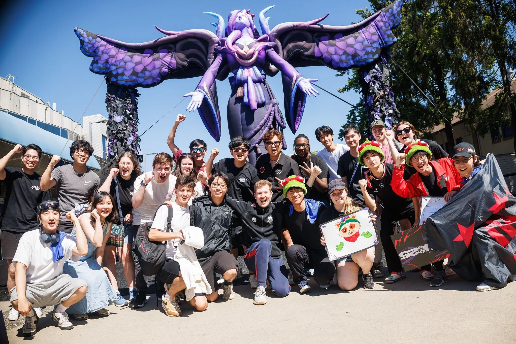 A group of young people posing for a photo outside the LTA (formerly LCS) Championship event, with a large purple-and-pink Morgana inflatable from League of Legends in the background. The group appears excited, with some cheering and flexing their mu