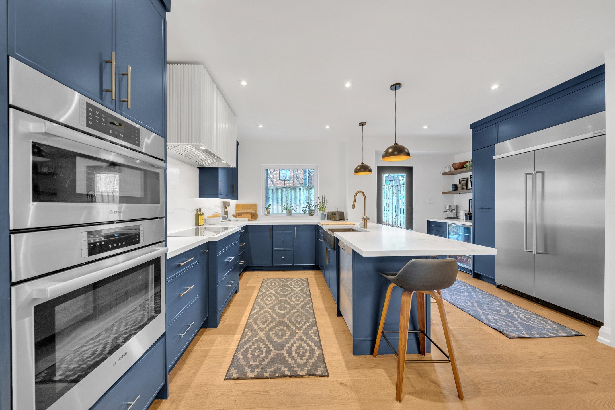 This modern Boho kitchen features vibrant blue cabinets and light wood tones, accented with sleek, stainless steel appliances. A white countertop island provides extra workspace and seating, complemented by stylish stools. Patterned rugs and industri