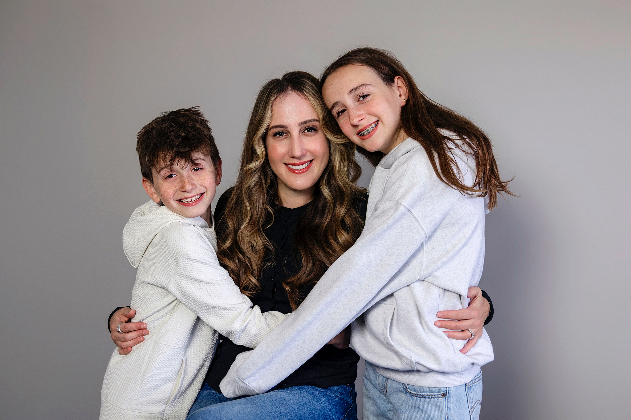 A woman hugging two children, a boy and a girl, against a plain gray background during mommy and me mini session in Northville, Michigan.