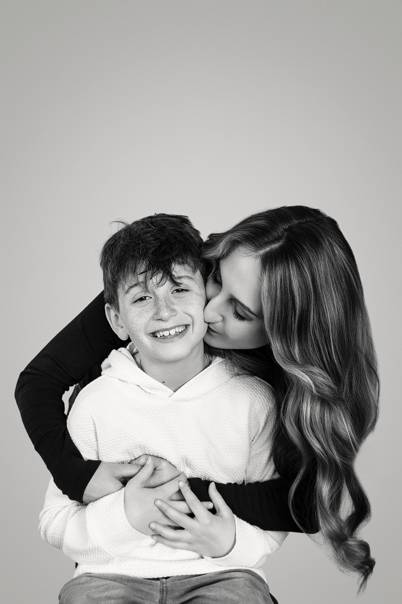 A black and white photo of a woman with long, wavy hair kissing a smiling young boy on the cheek.
