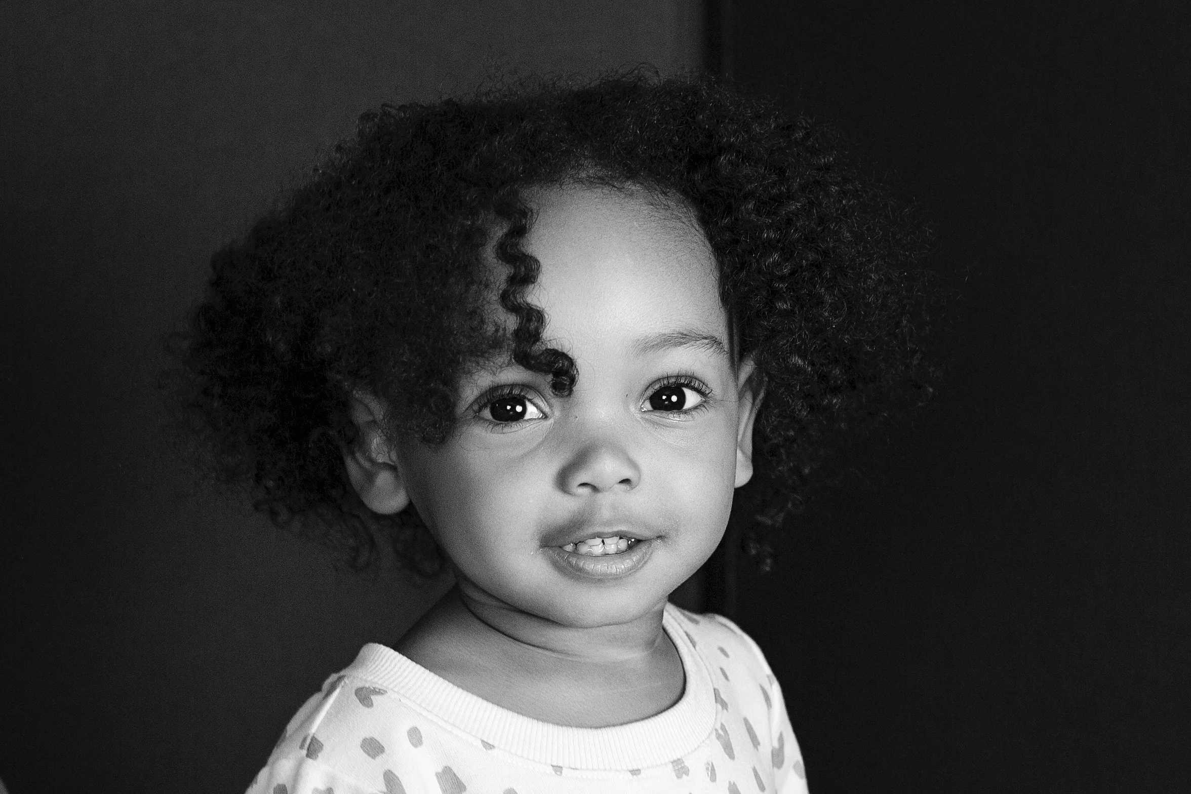 A young girl with curly hair and big eyes, smiling slightly, wearing a light-colored shirt with a pattern of small shapes, against a dark background.