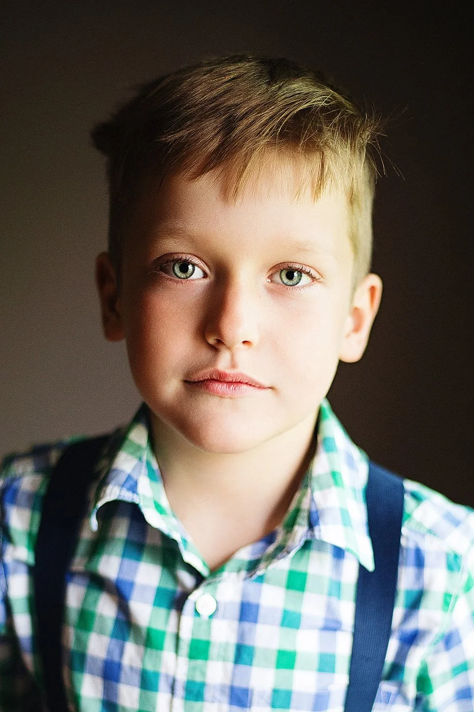 Close-up of a young boy with blond hair and blue eyes, wearing a checkered green, blue, and white shirt with suspenders, looking directly at the camera.