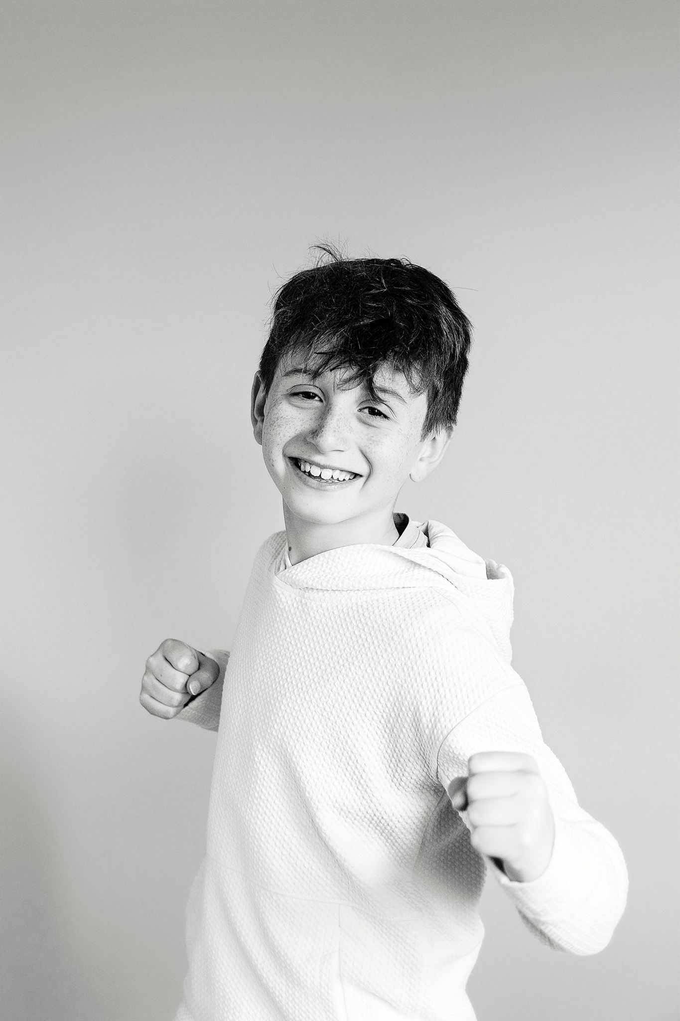 A young boy with short, tousled dark hair, smiling and making a fist with both hands in a fighting stance, wearing a textured white hoodie, against a plain light background during mommy and me mini session in Northville, Michigan.