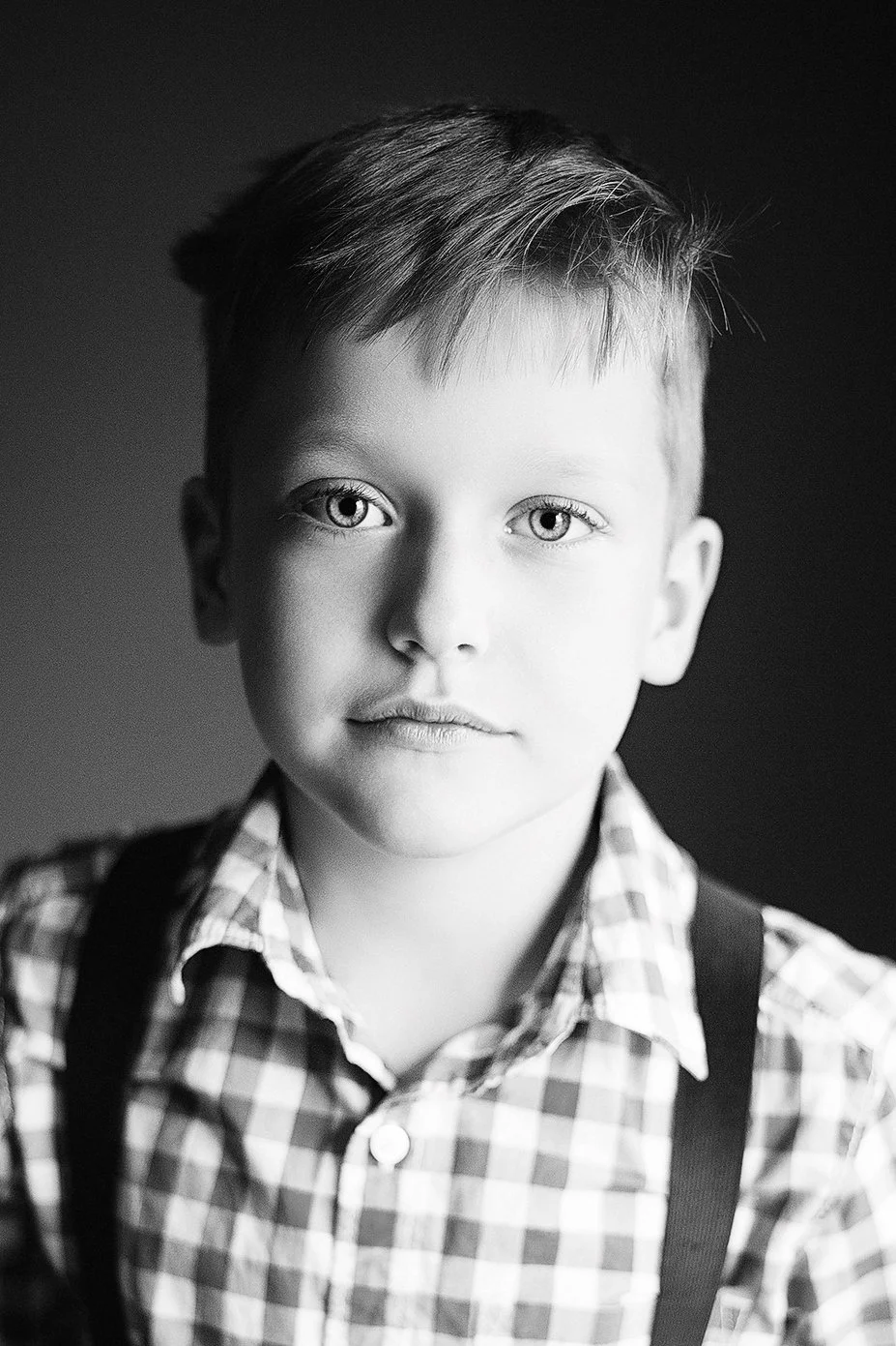 Black and white portrait of a young boy with short hair, wearing a checkered shirt and suspenders, looking directly at the camera.
