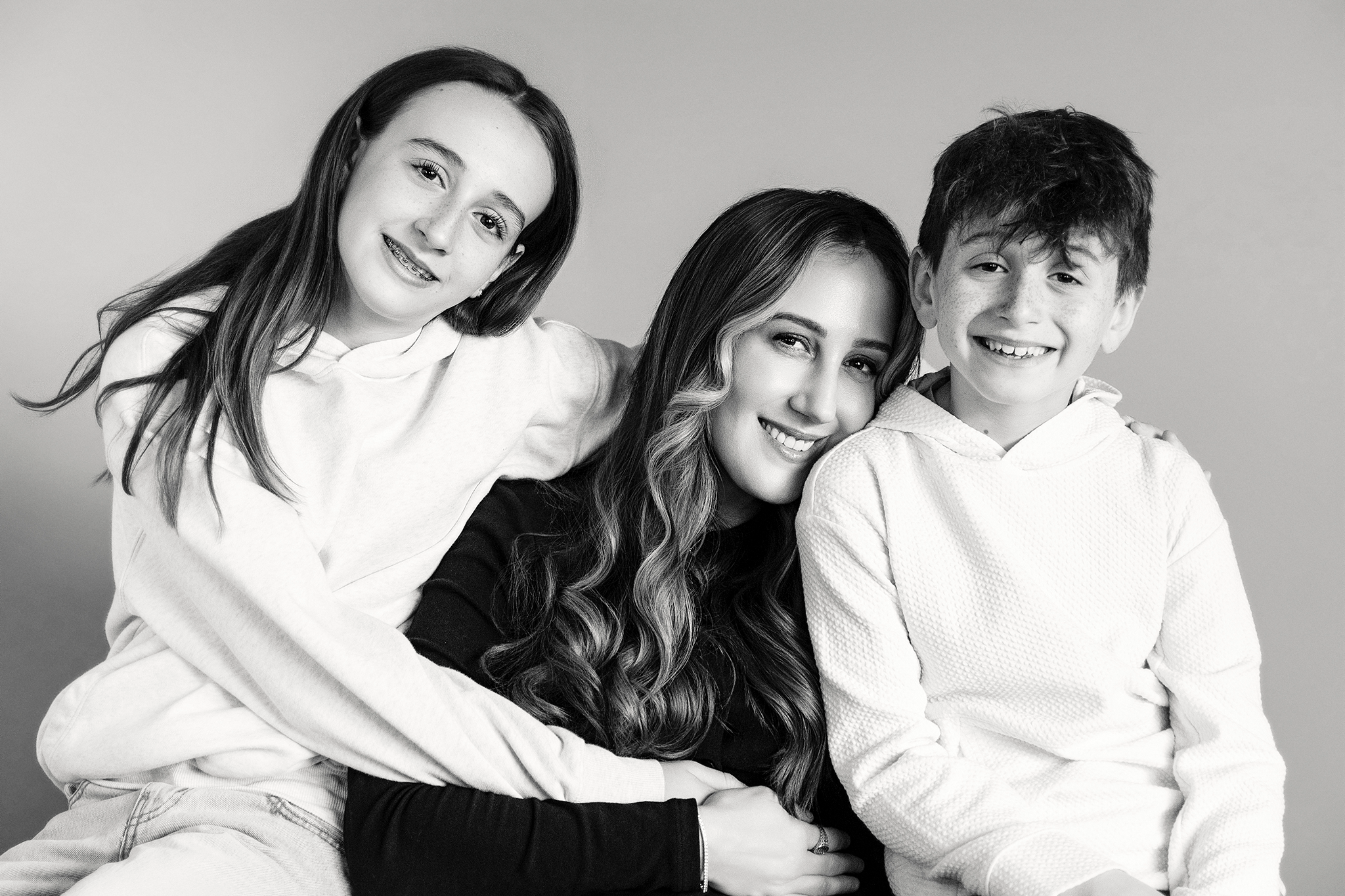 Three people, a woman, a young girl and a boy, sitting close together, smiling during mommy and me mini session in Northville, Michigan, in a black and white photograph.