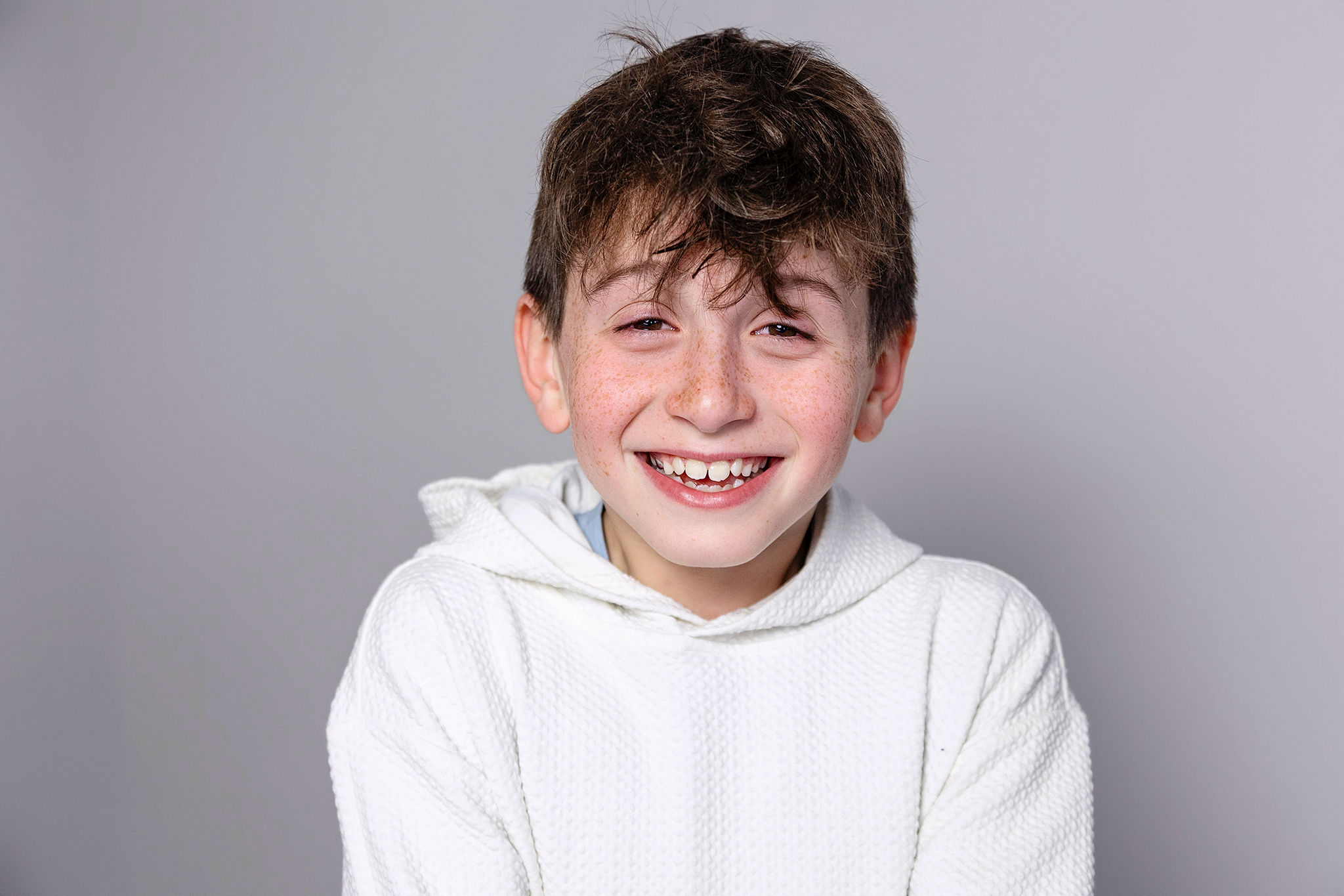 A smiling young boy with brown hair and freckles, wearing a white textured hoodie, standing against a plain gray background during mommy and me mini session in Northville, Michigan.