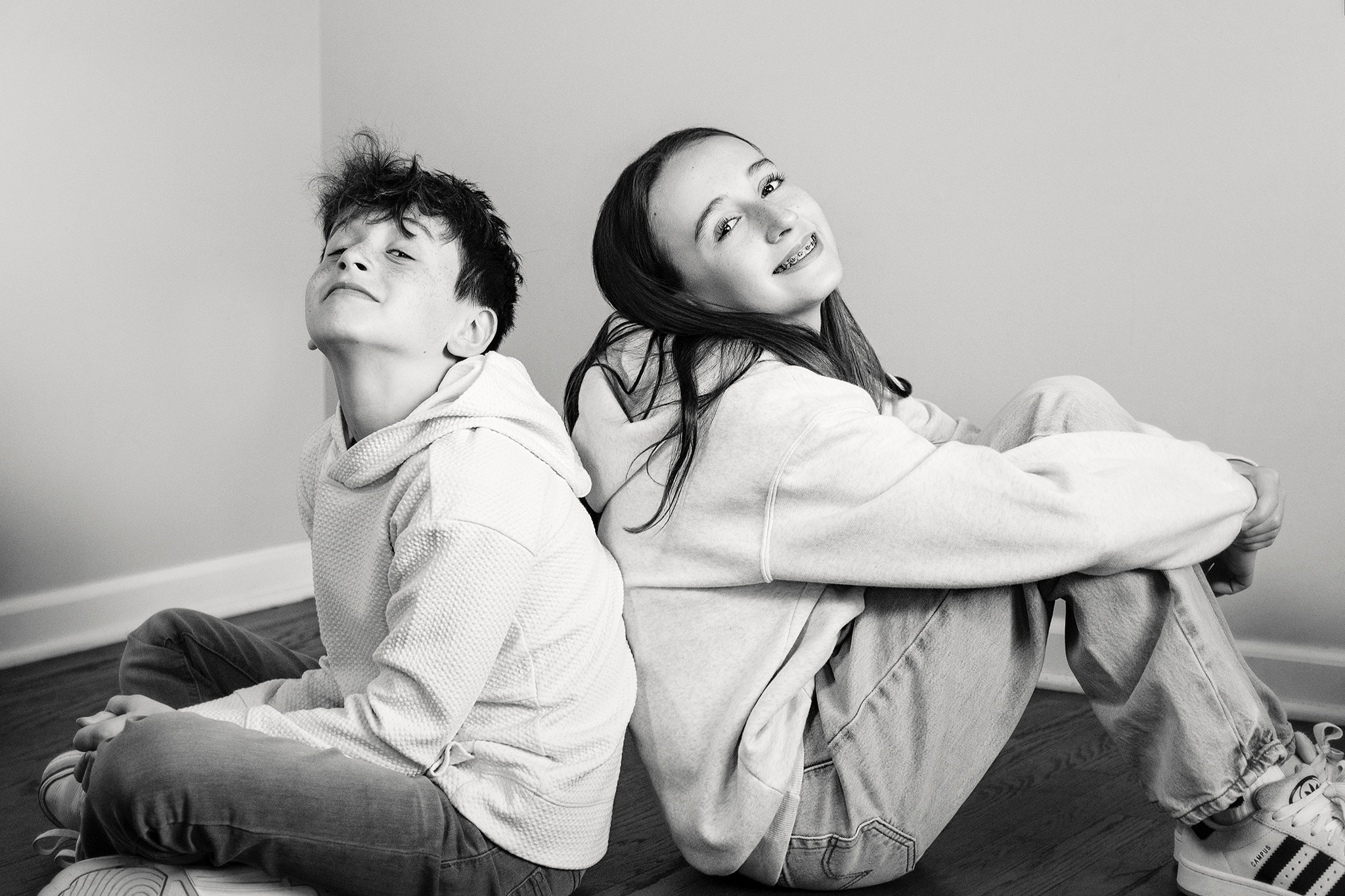 Two teenagers sitting on the floor, back-to-back, smiling and looking at the camera, in a minimalistic room with plain walls during mommy and me mini session in Northville, Michigan.