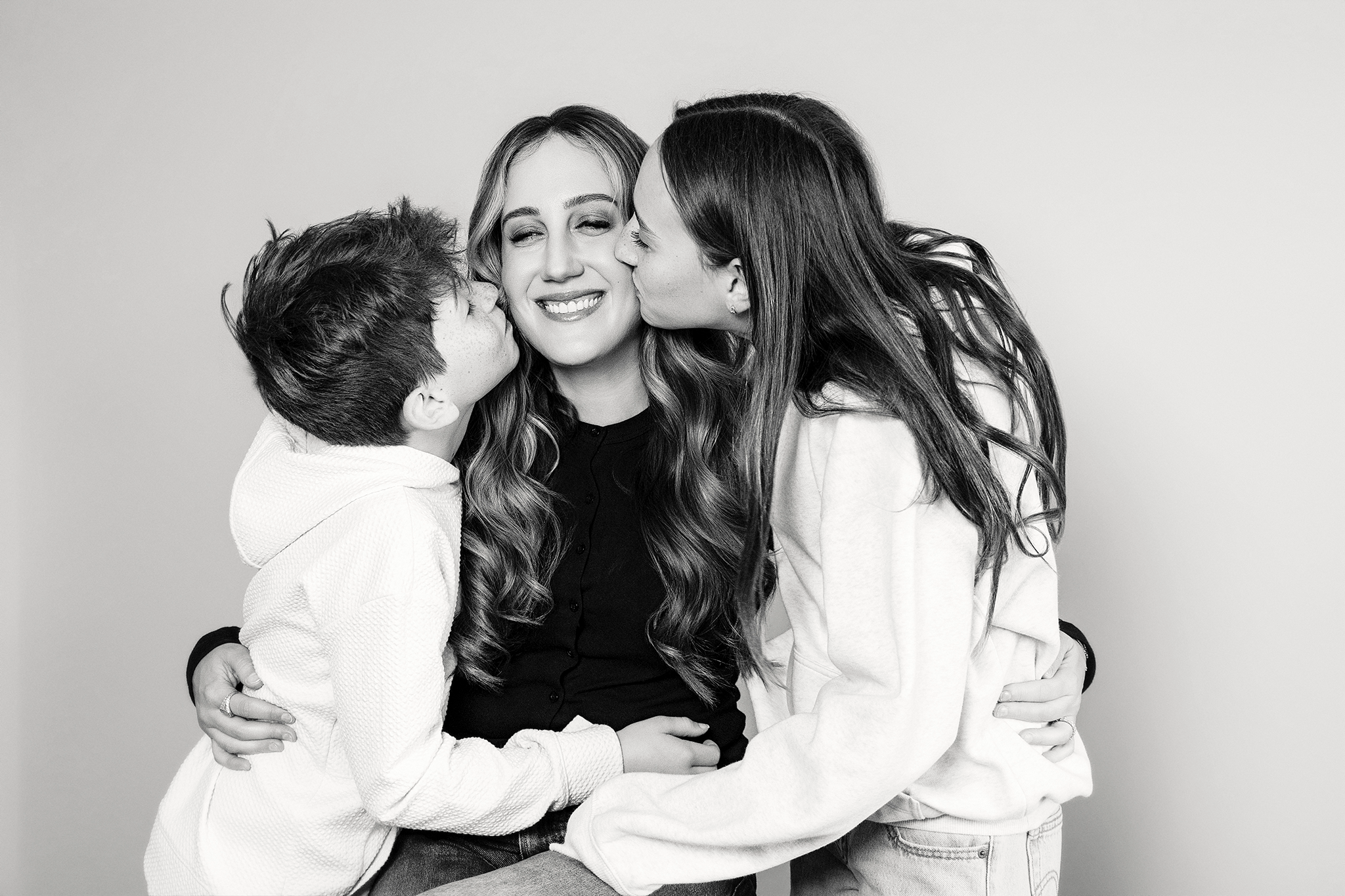 A family of three hugging during mommy and me mini session in Northville, Michigan, two children kissing the woman in the middle on the cheeks in black and white.