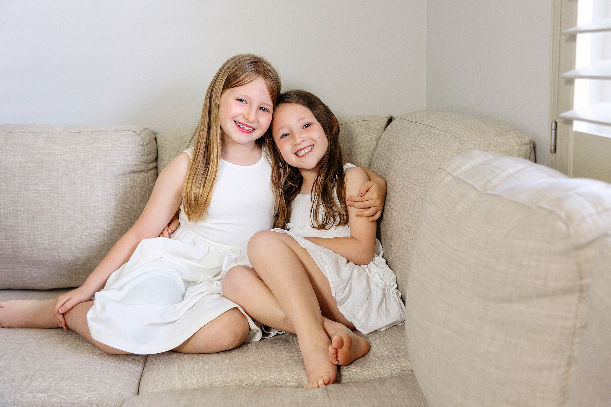 Two young girls with long hair sitting on a beige couch, smiling during mommy and me mini session in Northville, Michigan, with one girl hugging the other; background includes a white wall and a window with blinds.