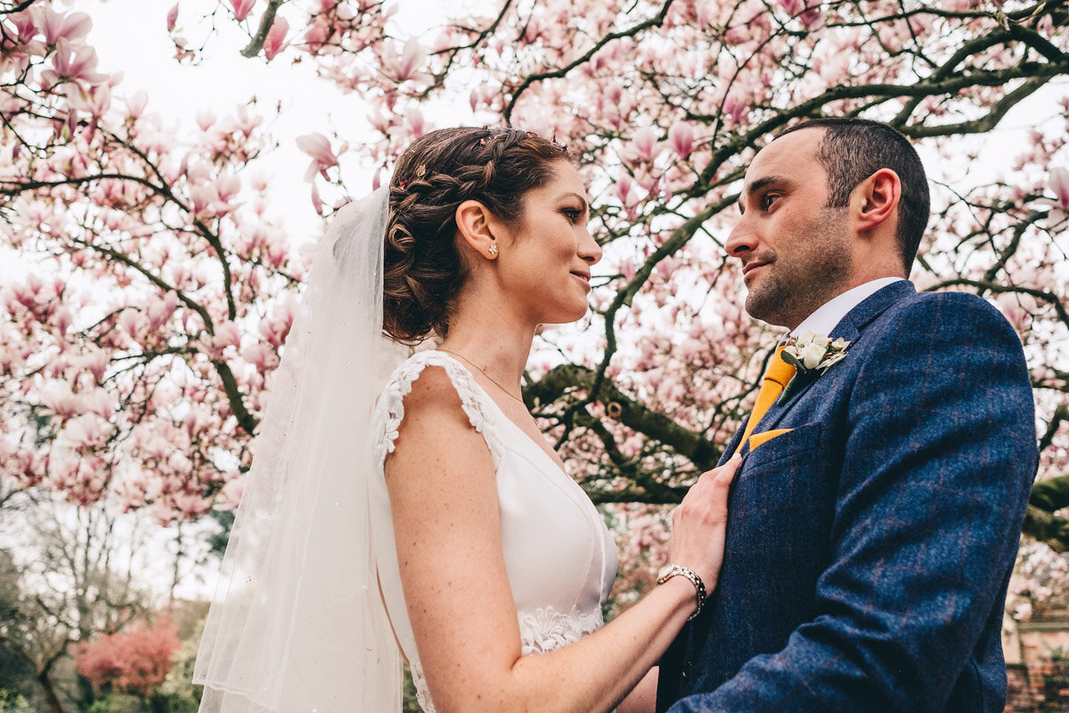 Bride and groom stand under blossoming tree in Didsbury Parsonage Gardens