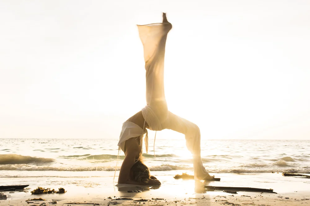 A woman in a wide-legged headstand at the water&rsquo;s edge, backlit by the sun.