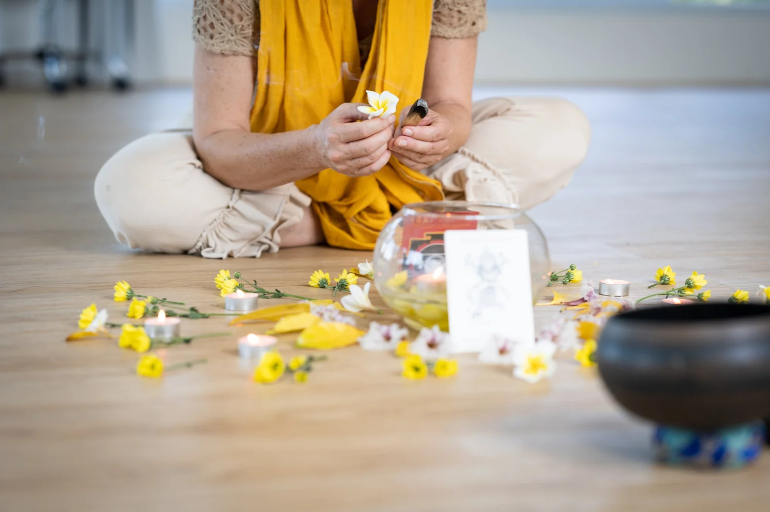 A woman sits cross-legged on a wooden floor, holding a white flower above an offering of yellow flowers, tea lights and a glass bowl, with a singing bowl in front.