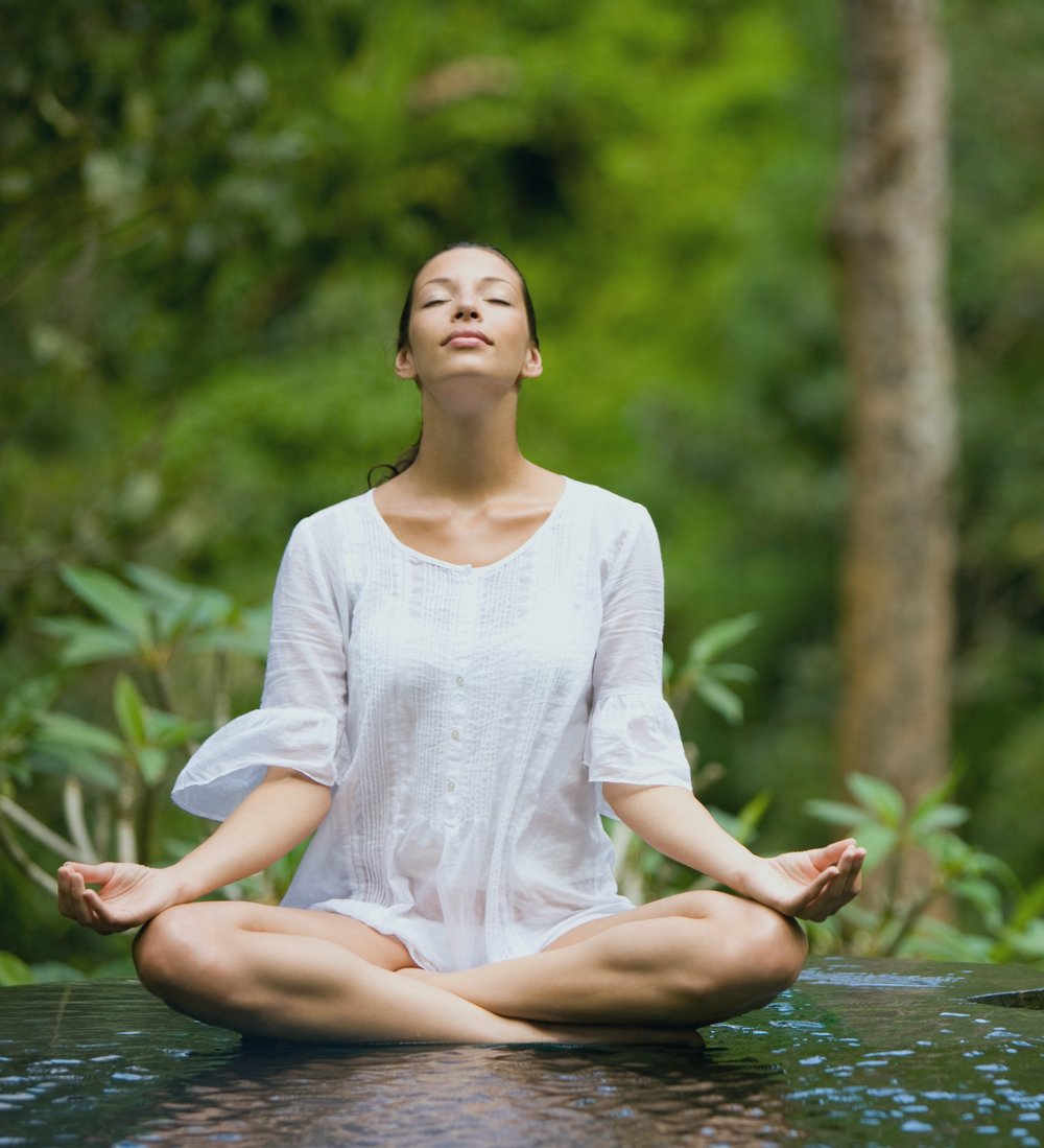 A woman in white meditating cross-legged above water, eyes closed, jungle behind.