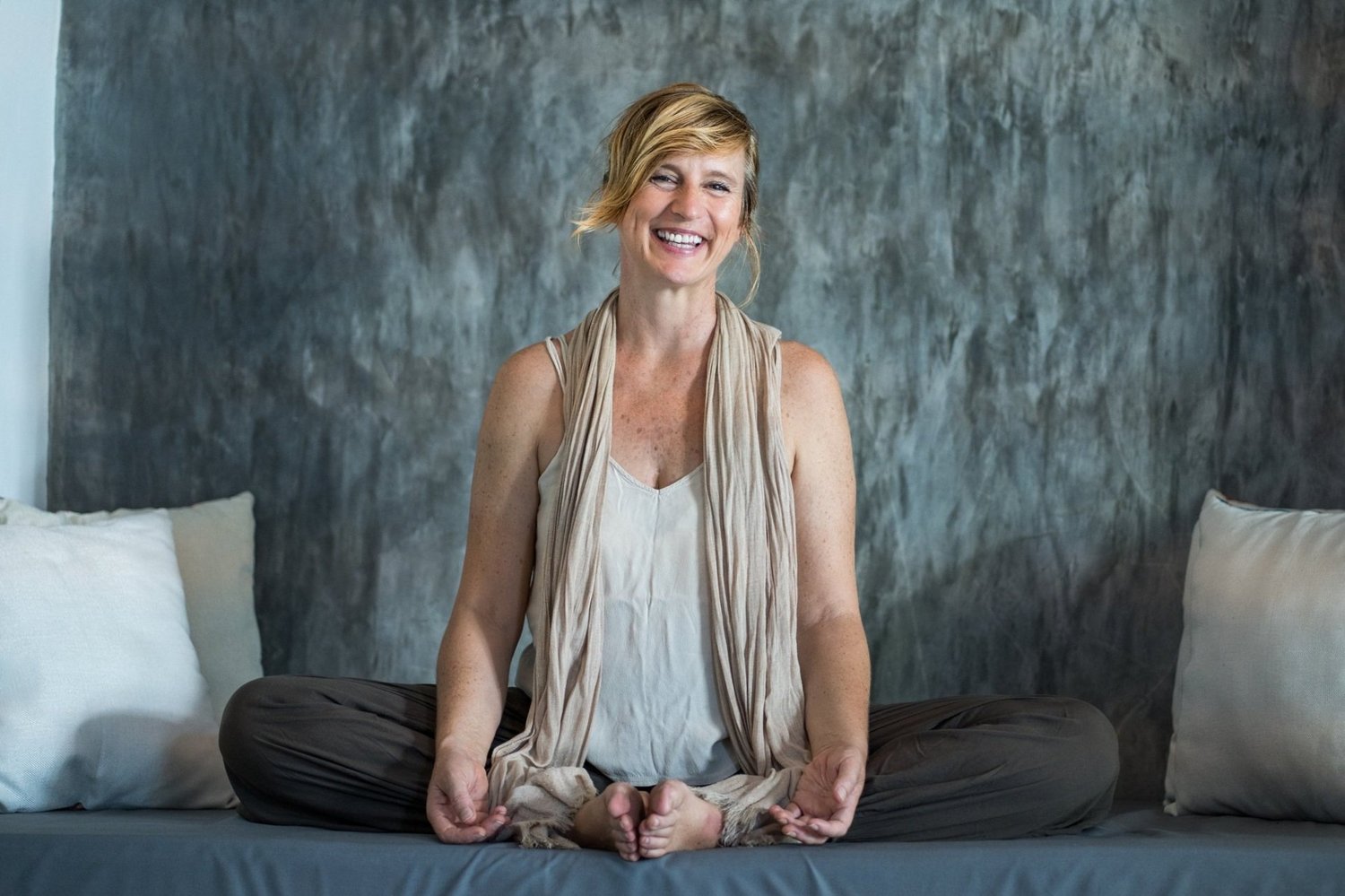 Lorraine Taylor, smiling, sits on a grey daybed with the soles of her feet together.