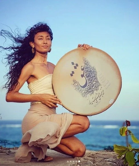 Dominique Zuniga kneeling by the sea, holding a frame drum.