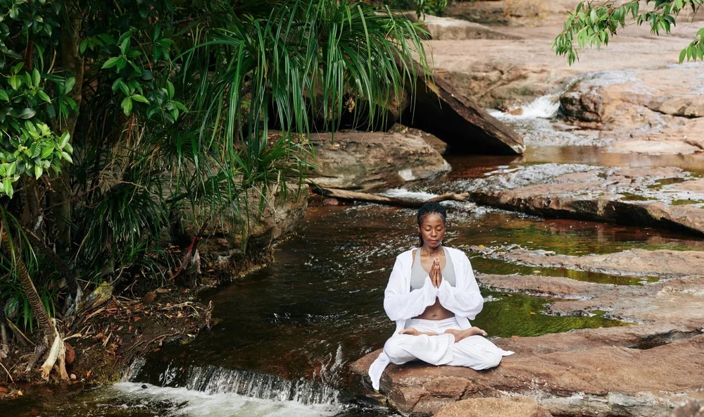 A woman in white meditating on rocks beside a small waterfall.