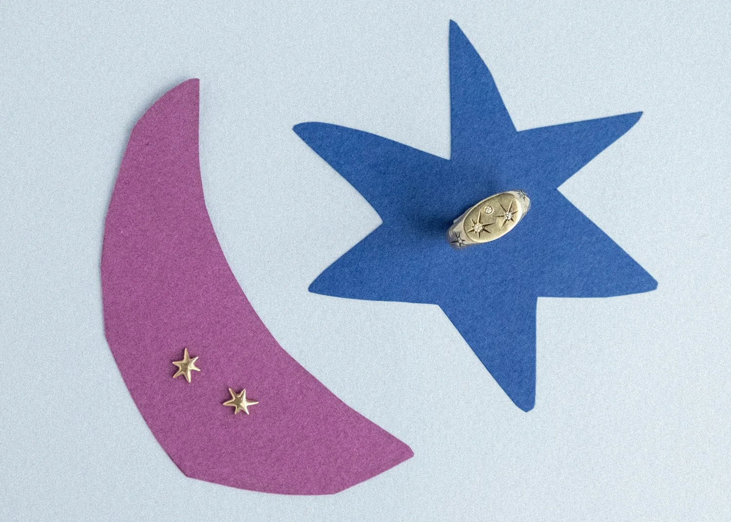 Gold star-shaped earrings and a gold signet ring on top of colorful cut-outs of a moon and a star