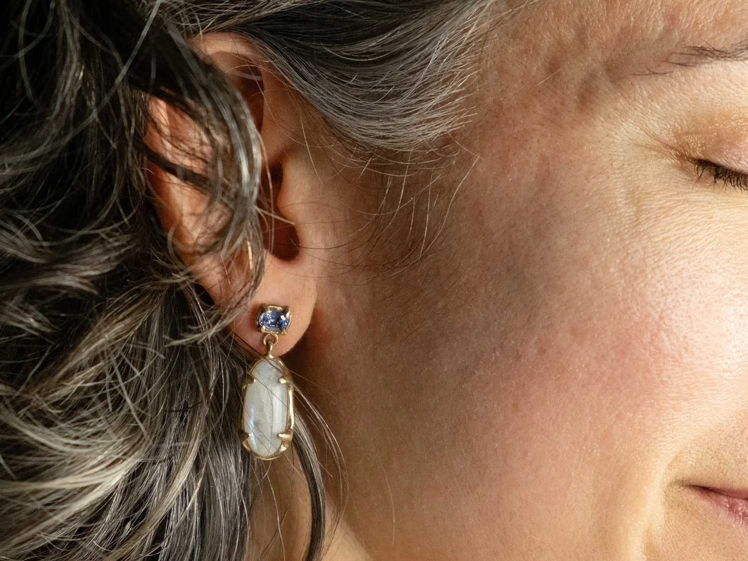 Closeup of female model wearing 14k gold earrings with top sapphire stone and hanging moonstone