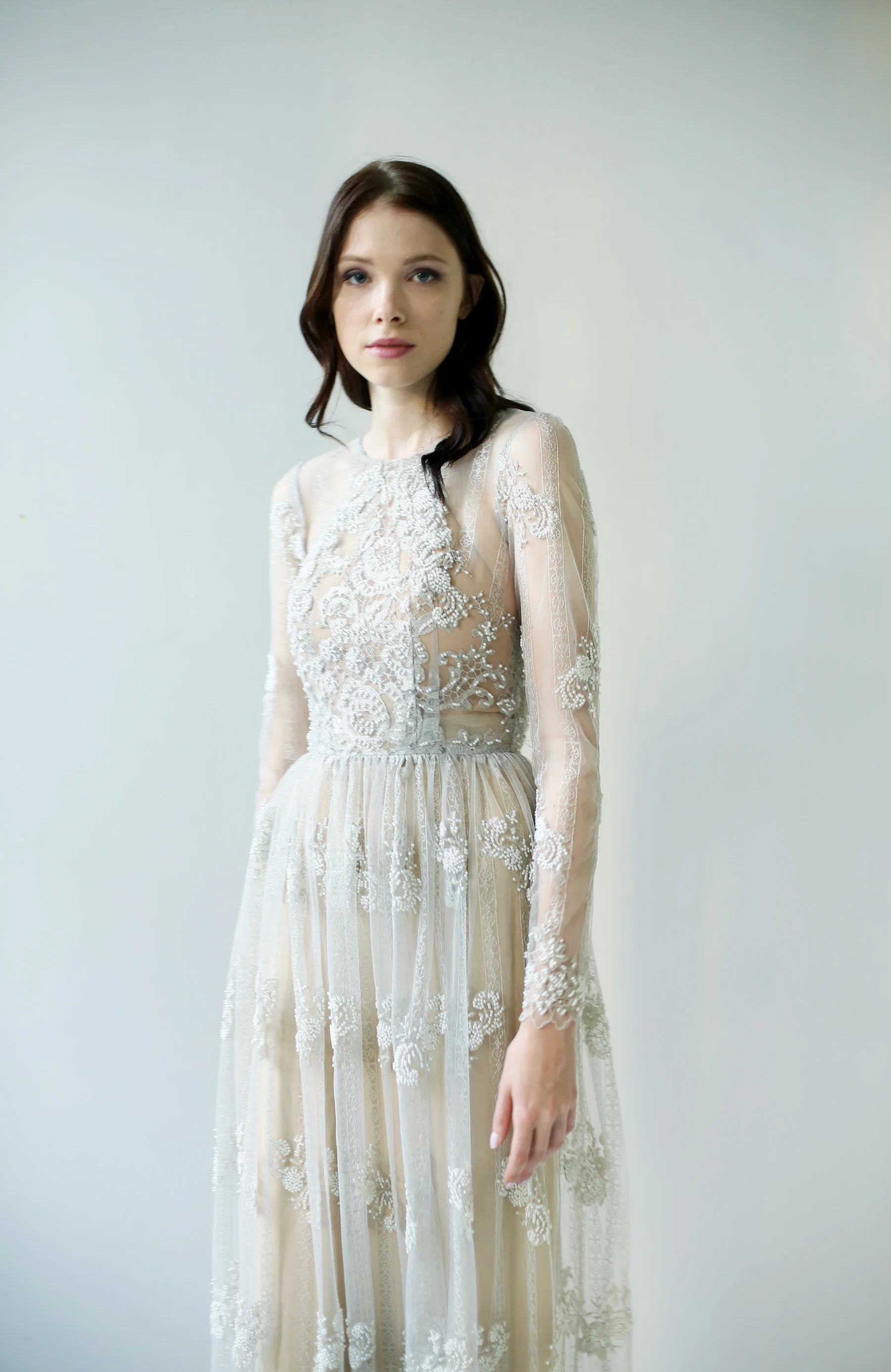 long sleeved, gray beaded gown with ornately embellished bodice and hem. The skirt is a softly gathered column with inset train. It is layered over a nude halter, backless slipdress.