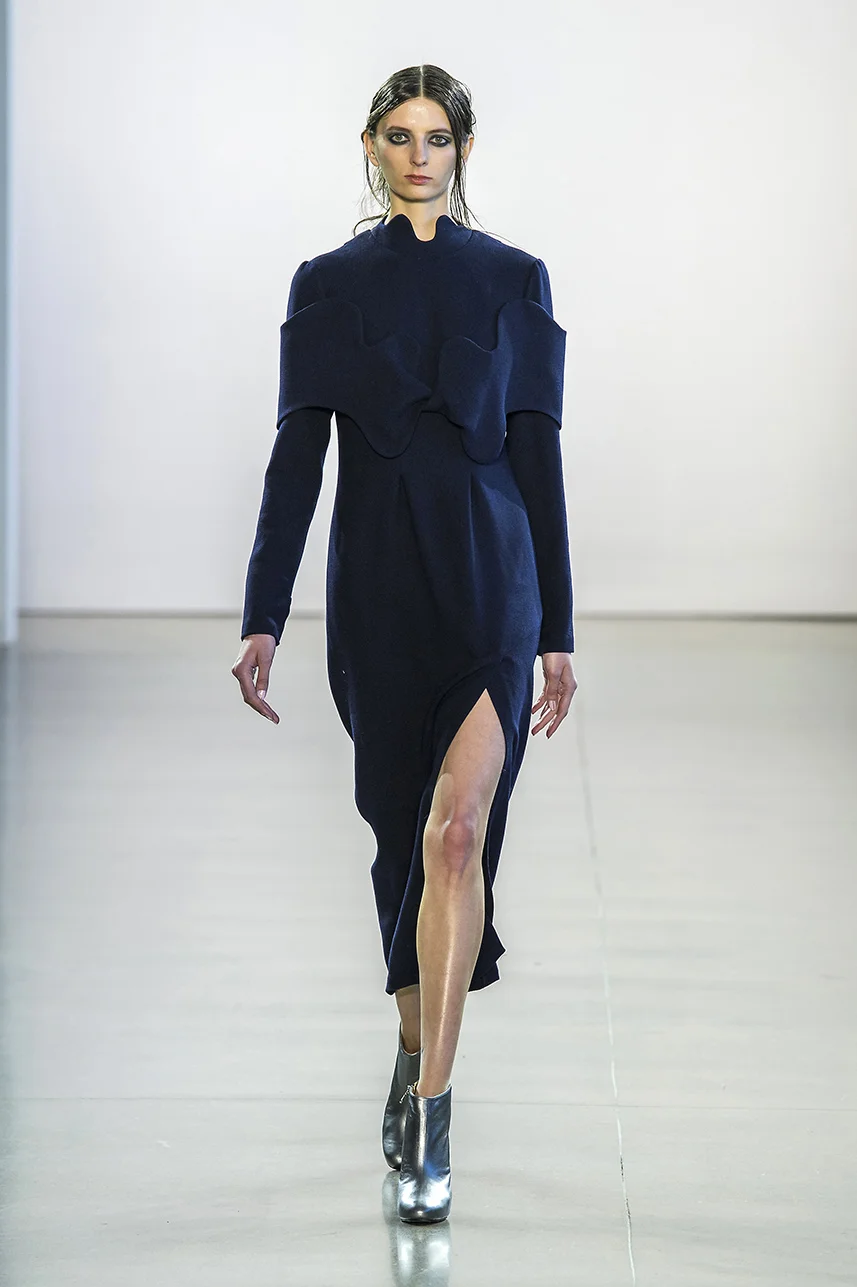 Navy wool crepe dress with high neck and side slit. , off-shoulder architectural detail. Slim cut silhouette shaped with darts, with long sleeves and high neckline. Dress zips up the back.