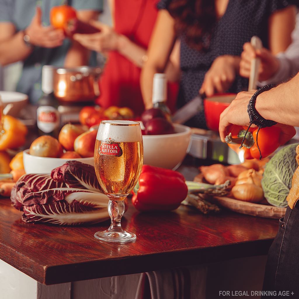 Cook with Friends, Stella Artois. People gathered in a kitchen with glasses of beer and lots of produce.