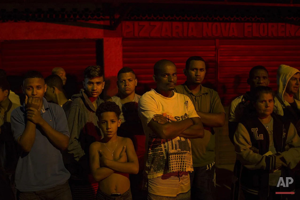 In this July 15, 2016 photo, residents watch as police work the crime scene where a man was murdered in Mage, greater Rio de Janeiro, Brazil. Scenes of impunity and violence play out daily in many of Rio's hundreds of slums, known here as favelas, and other outlying areas. The vast majority of killings are the result of heavily armed gangs who frequently shoot it out in turf wars. (AP Photo/Felipe Dana)