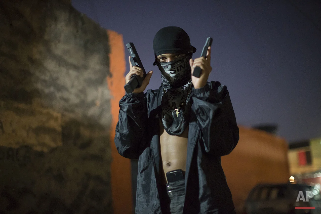 In this July 11, 2016 photo, a young, masked drug trafficker poses for photos holding his guns at a slum in Rio de Janeiro, Brazil. Teenagers openly tote guns while they work as guards, lookouts and distributors for drug lords operating just a few miles (kilometers) from where hundreds of thousands and tourists and athletes will be for the Aug. 5-21 Olympic Games. (AP Photo/Felipe Dana)