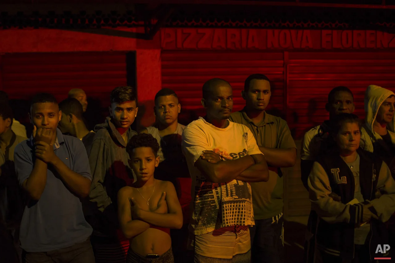 In this July 15, 2016 photo, residents watch as police work the crime scene where a man was murdered in Mage, greater Rio de Janeiro, Brazil. Scenes of impunity and violence play out daily in many of Rio's hundreds of slums, known here as favelas, and other outlying areas. The vast majority of killings are the result of heavily armed gangs who frequently shoot it out in turf wars. (AP Photo/Felipe Dana)