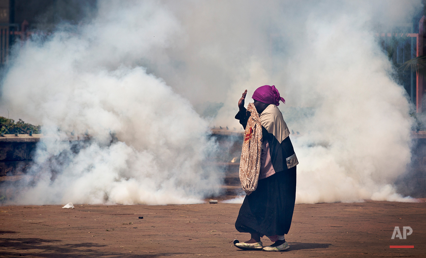  An elderly woman caught up in the clashes holds her hands in the air as a riot policeman approach amidst clouds of tear gas, during a protest in downtown Nairobi, Kenya Monday, May 16, 2016. Kenyan police have tear-gassed and beaten opposition suppo