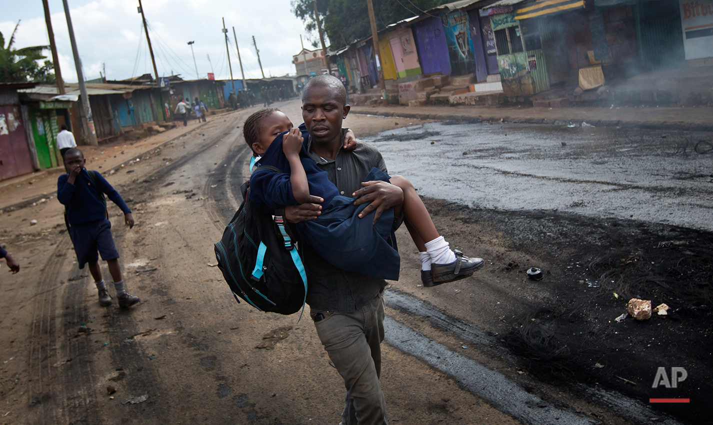  A man carries a schoolgirl overcome by tear gas to safety, past the charred remains of a burning barricade, as police firing tear gas engage protesters throwing rocks in the Kibera slum of Nairobi, Kenya Monday, May 23, 2016. Kenya's police shot, be