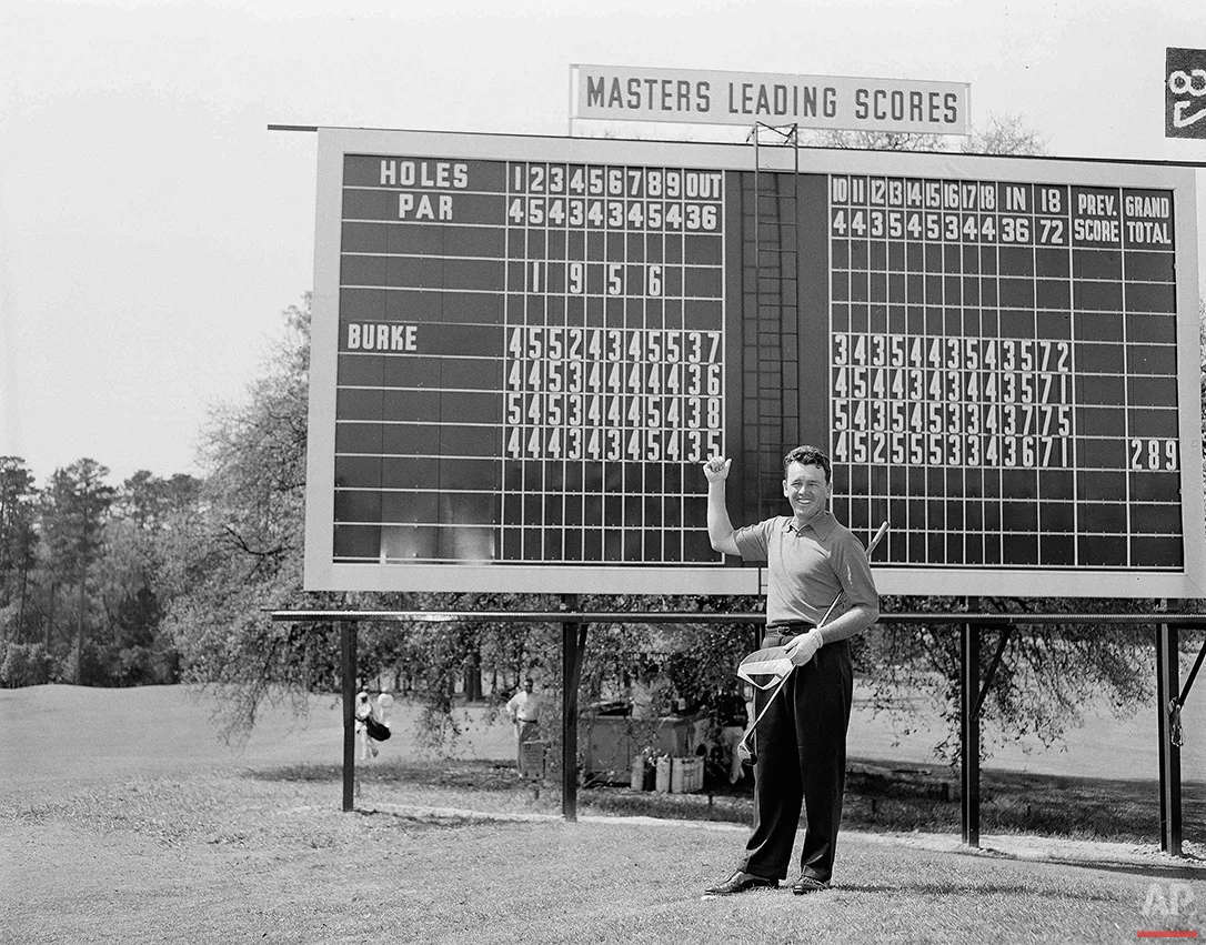 Defending Masters champ Jack Burke Jr. points a thumb to his 1956 winning round scores which are posted along the No. 1 fairway of the Augusta National Golf Club, April 3, 1957. Burke surged from behind last year and overtook Ken Venturi on the last day with a total 289. (AP Photo/Horace Cort)
