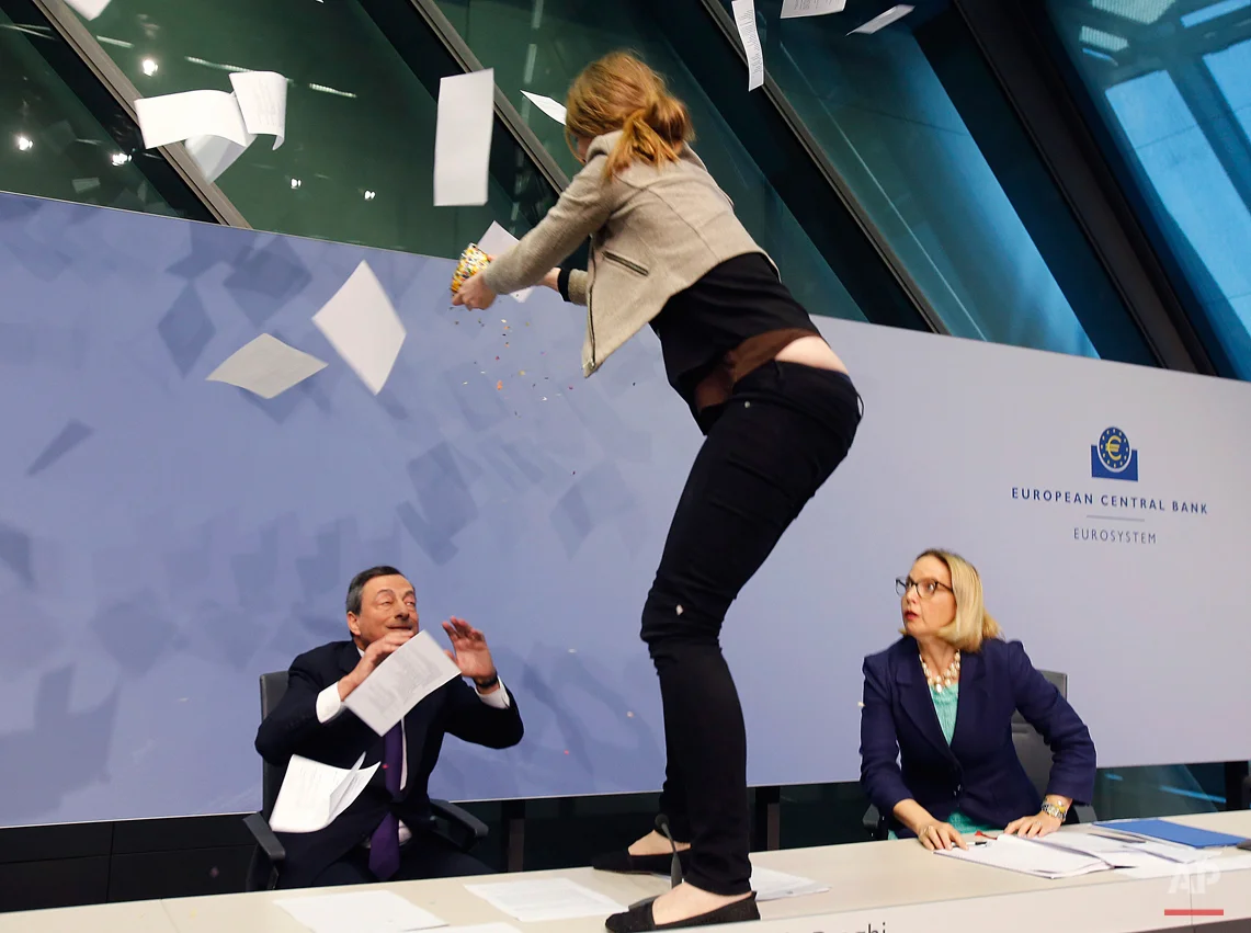 An activist stands on the table of the podium throwing paper at ECB President Mario Draghi, left, as Christine Graeff, Director General of Communications, looks on during a press conference of the European Central Bank, ECB, in Frankfurt, Germany, Wednesday, April 15, 2015. (AP Photo/Michael Probst)