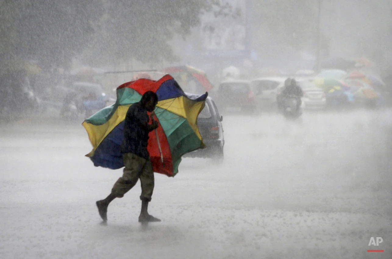 In this Friday, July 3, 2015, photo, a gust of wind bends the umbrella of a street vendor crossing the road during a heavy downpour in the eastern Indian city of Bhubaneswar, India. (AP Photo/Biswaranjan Rout)
