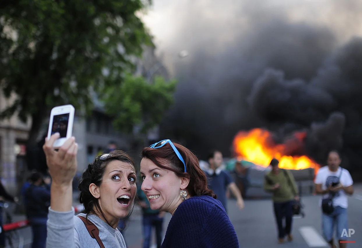 Tourists take a 'selfie' picture as demonstrators burn a trash container during a May Day rally in Barcelona, Spain, Thursday, May 1, 2014. Tens of thousands of workers marked May Day in European cities with a mix of anger and gloom over austerity measures imposed by leaders trying to contain the eurozone's intractable debt crisis. (AP Photo/Manu Fernandez)