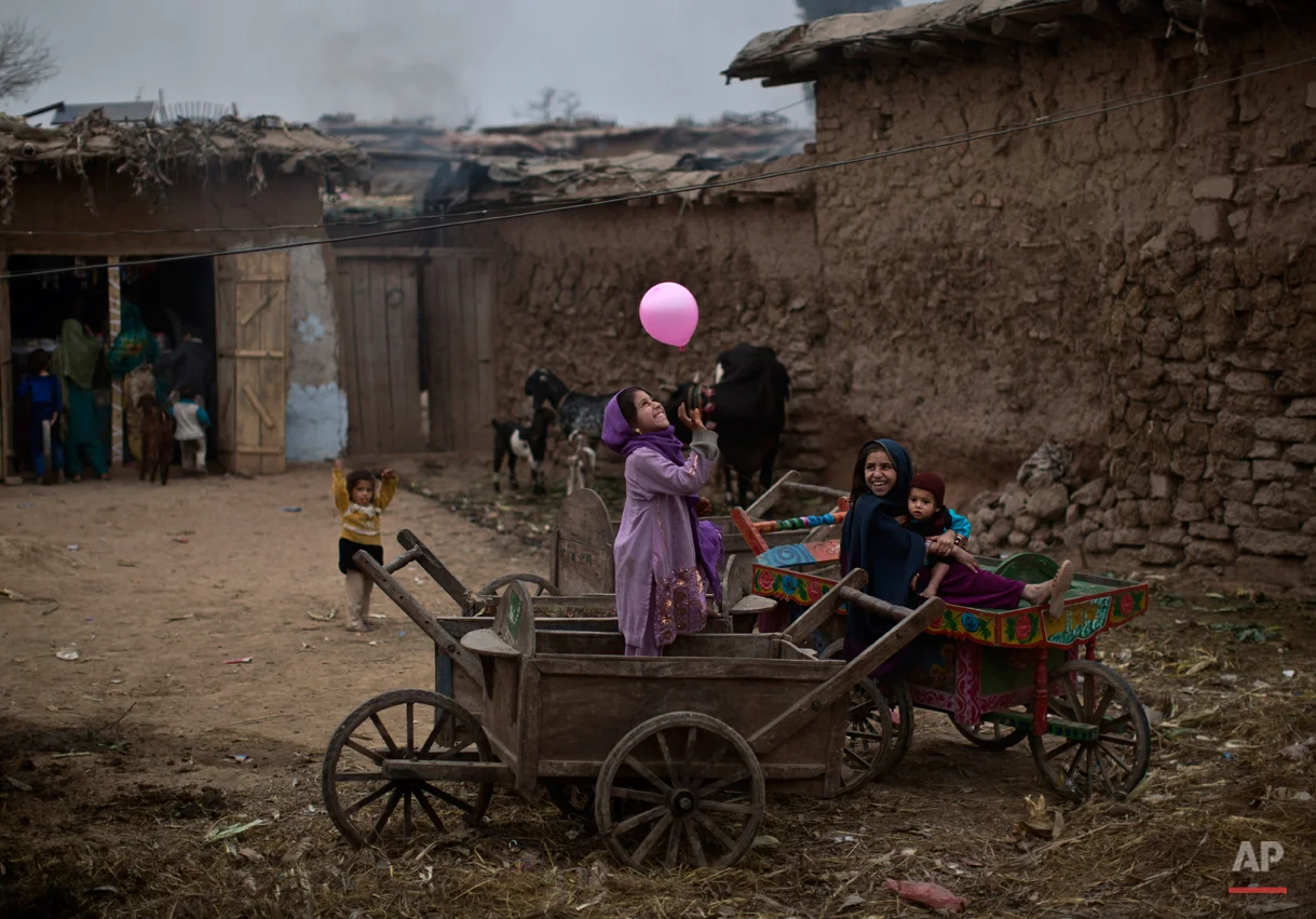 An Afghan refugee girl, right, holding her younger brother, sits on a wooden-cart looking at her friend playing with a balloon, in a poor neighborhood on the outskirts of Islamabad, Pakistan, Sunday, Feb. 2, 2014. (AP Photo/Muhammed Muheisen)